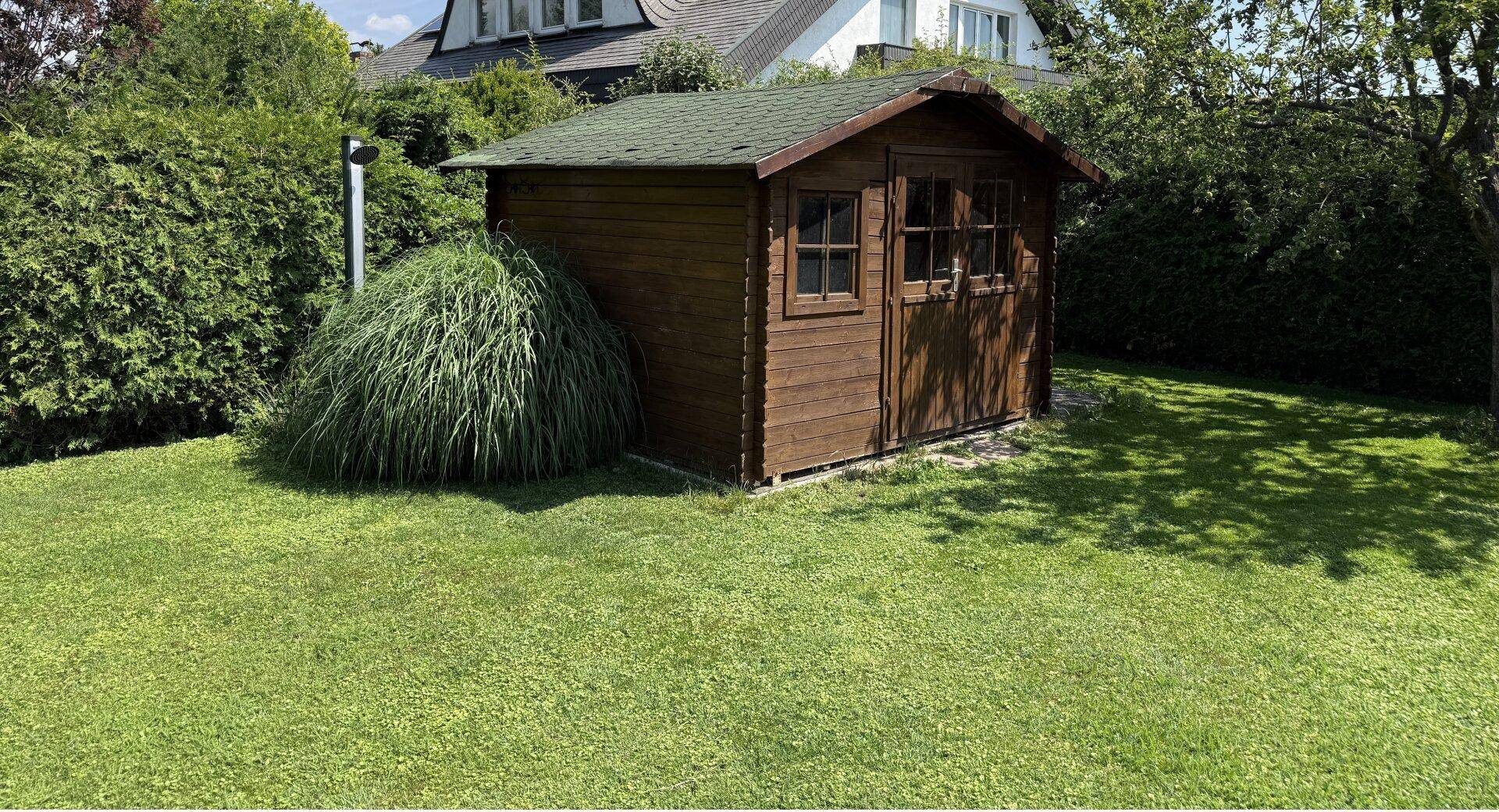 Kleines Holzgartenhaus auf dem gepflegten Rasen, umgeben von Grünpflanzen.