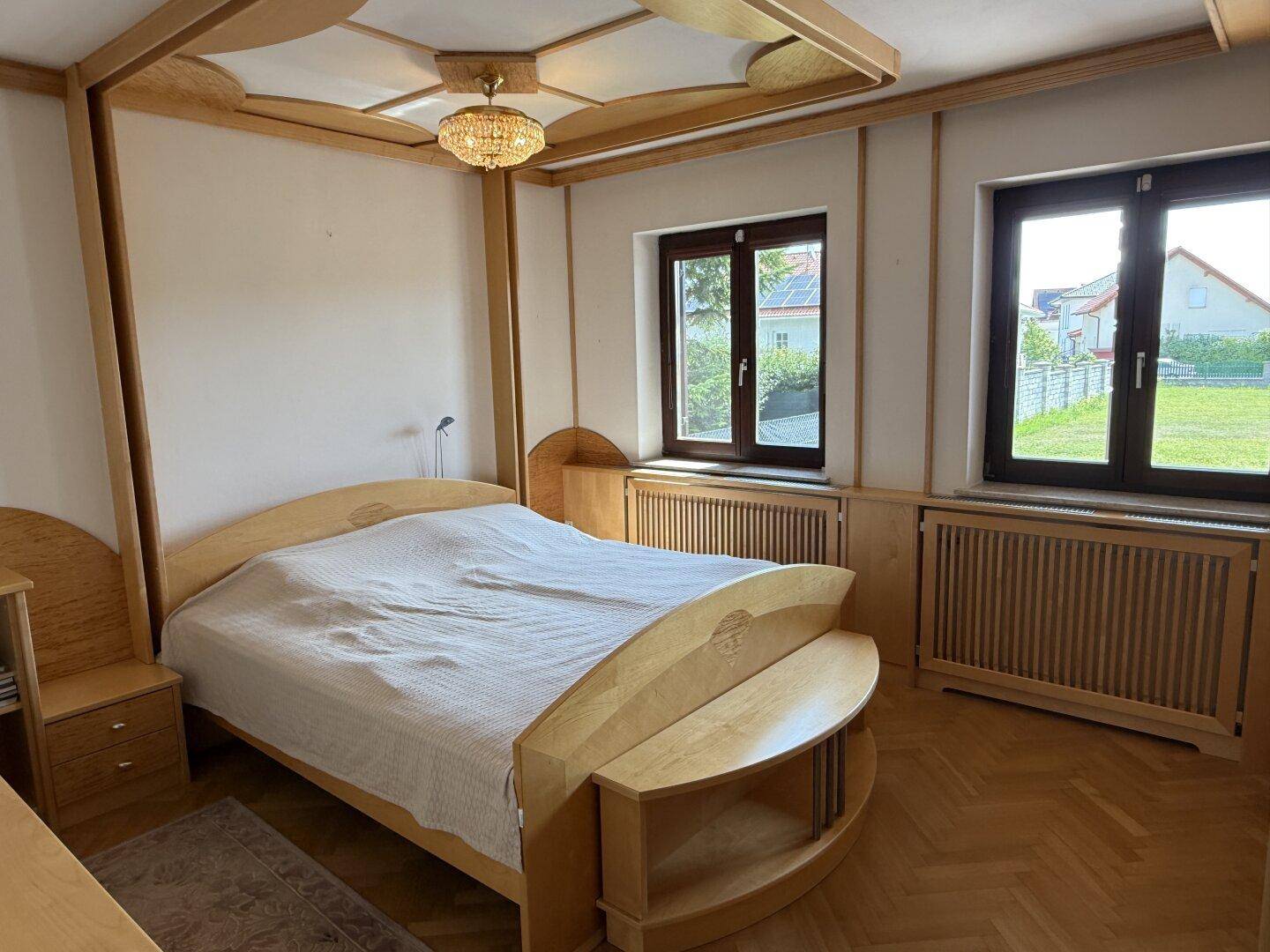 Helles Schlafzimmer mit Holzbett, Parkettboden und Fenstern mit Blick ins Grüne.