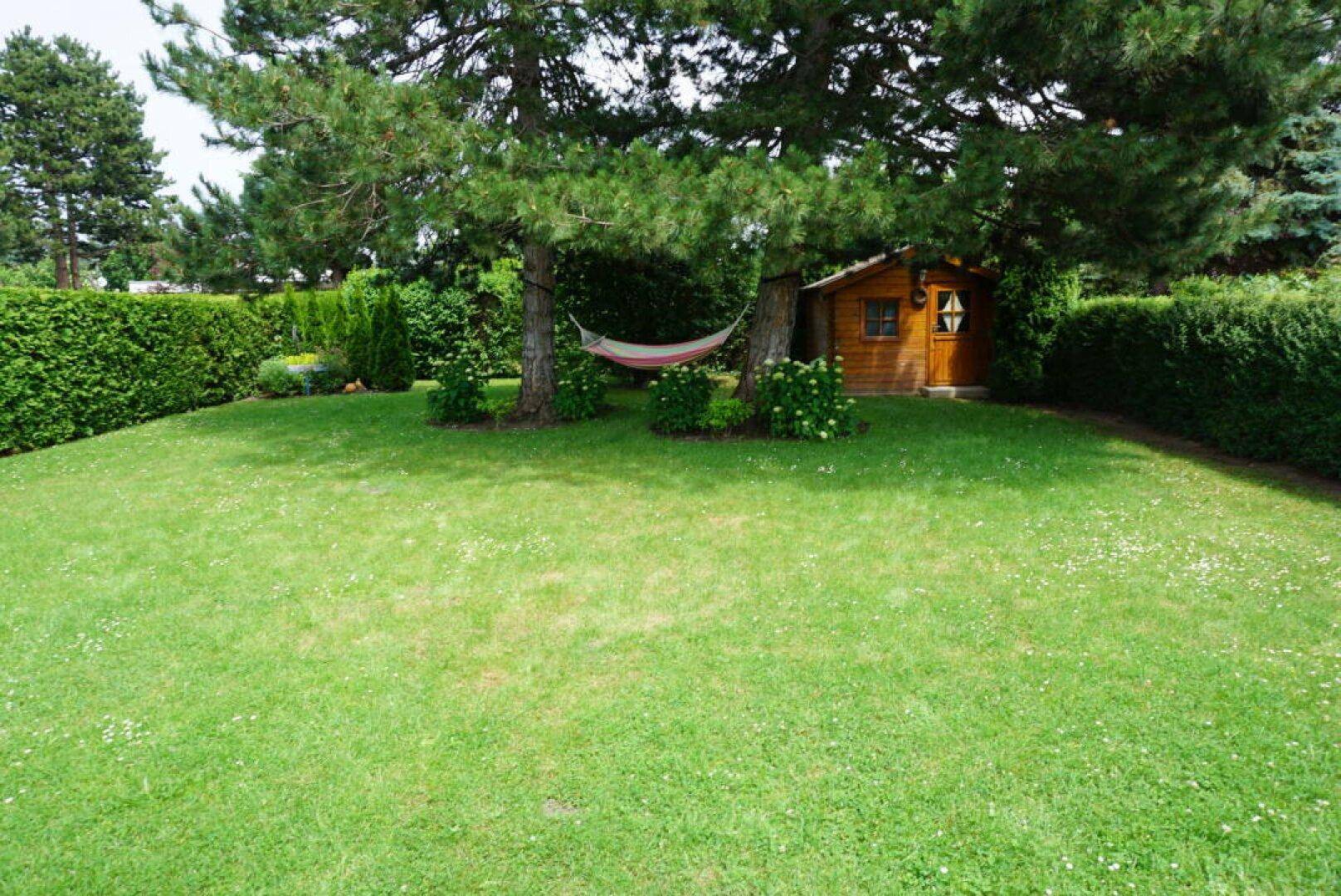 Gepflegter Garten mit Rasenfläche, Bäumen, Hängematte und einem kleinen Gartenhaus.
