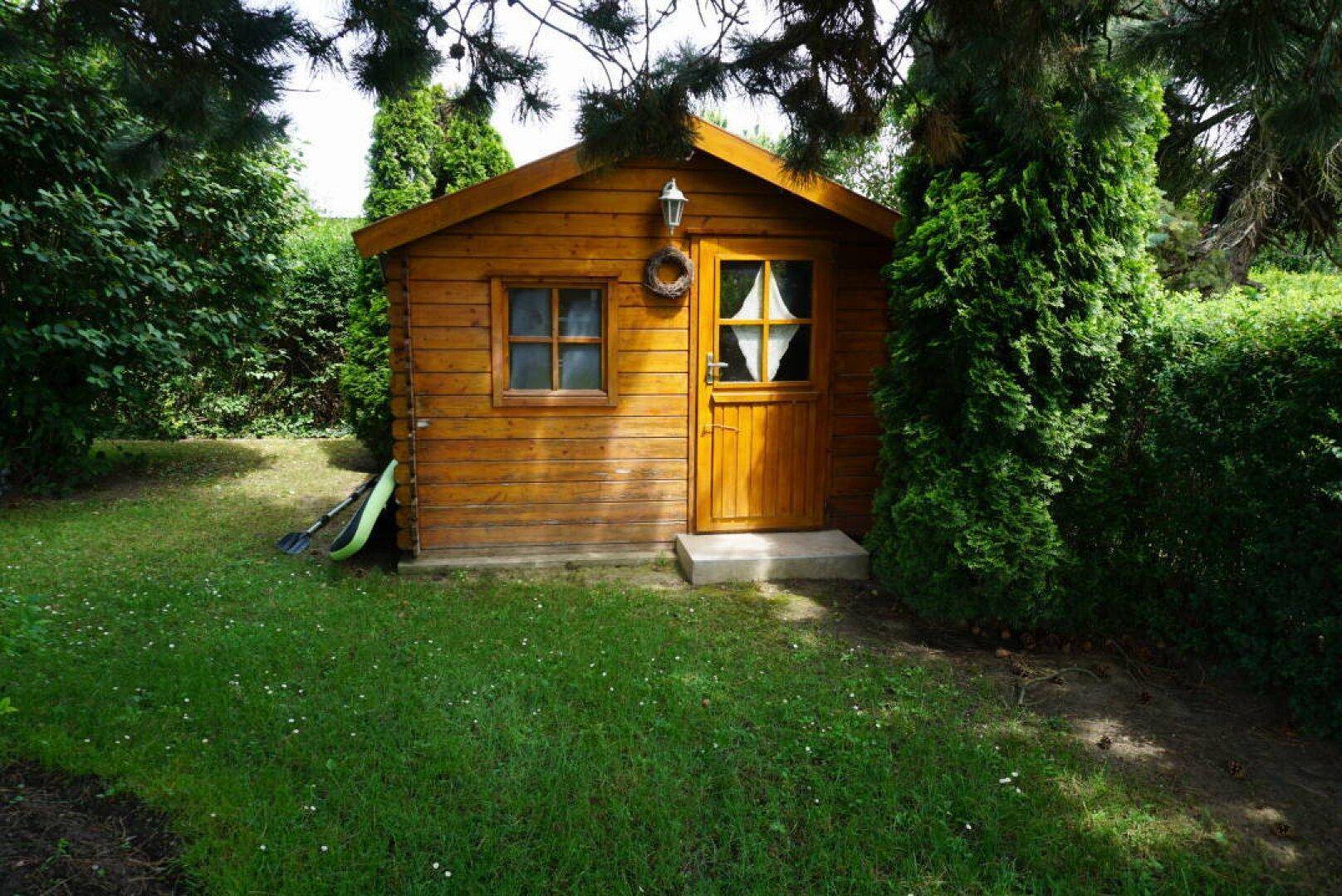 Rustikales Gartenhaus aus Holz, umgeben von grünen Hecken und Rasen.