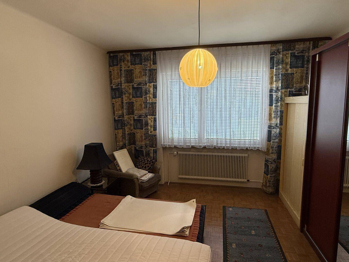 Schlafzimmer mit Doppelbett, Kleiderschrank und großem Fenster mit Jalousien.