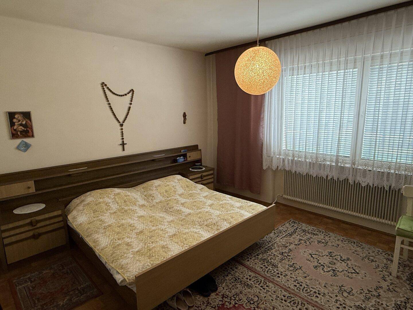 Schlafzimmer mit großem Doppelbett, Teppich und Fenster mit weißer Gardine.