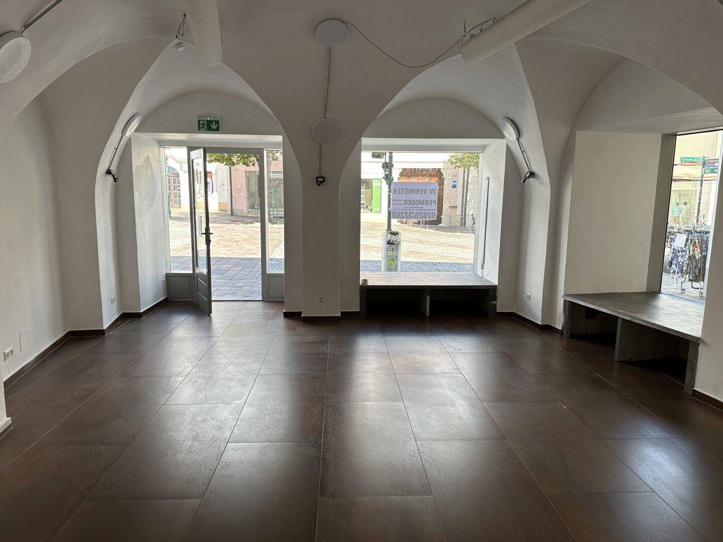 Heller Ladenbereich mit hohen Gewölbedecken, großen Schaufenstern zur Straße und dunklem Fliesenboden.