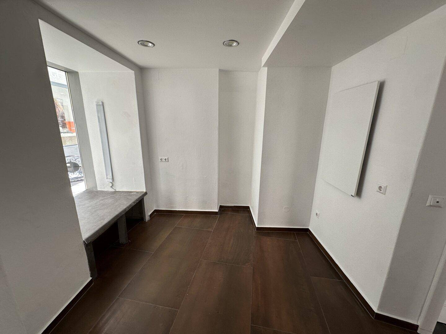 Schlichter Raum mit weißen Wänden, dunklem Fliesenboden und einem Fenster, das Tageslicht hereinlässt.