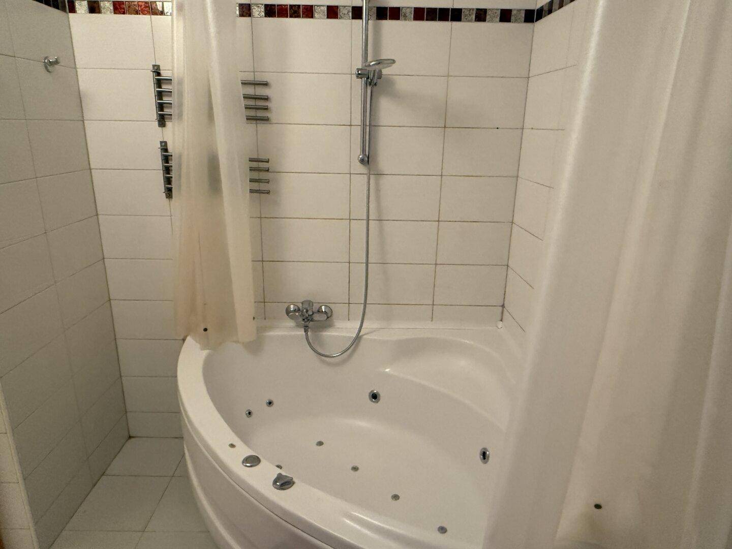 Badezimmer mit Eck-Whirlpool und Duschvorhang, komplett gefliest in Weiß mit dekorativer Bordüre.