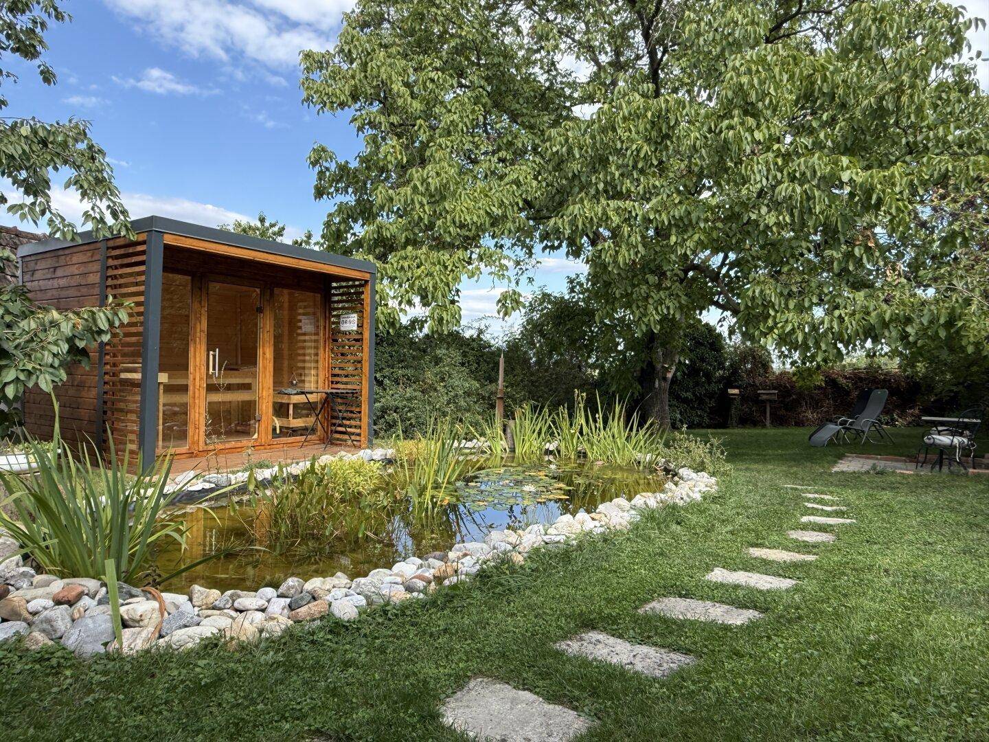 Weitläufiger Garten mit moderner Sauna, Teich und Liegestühlen unter schattenspendenden Bäumen.
