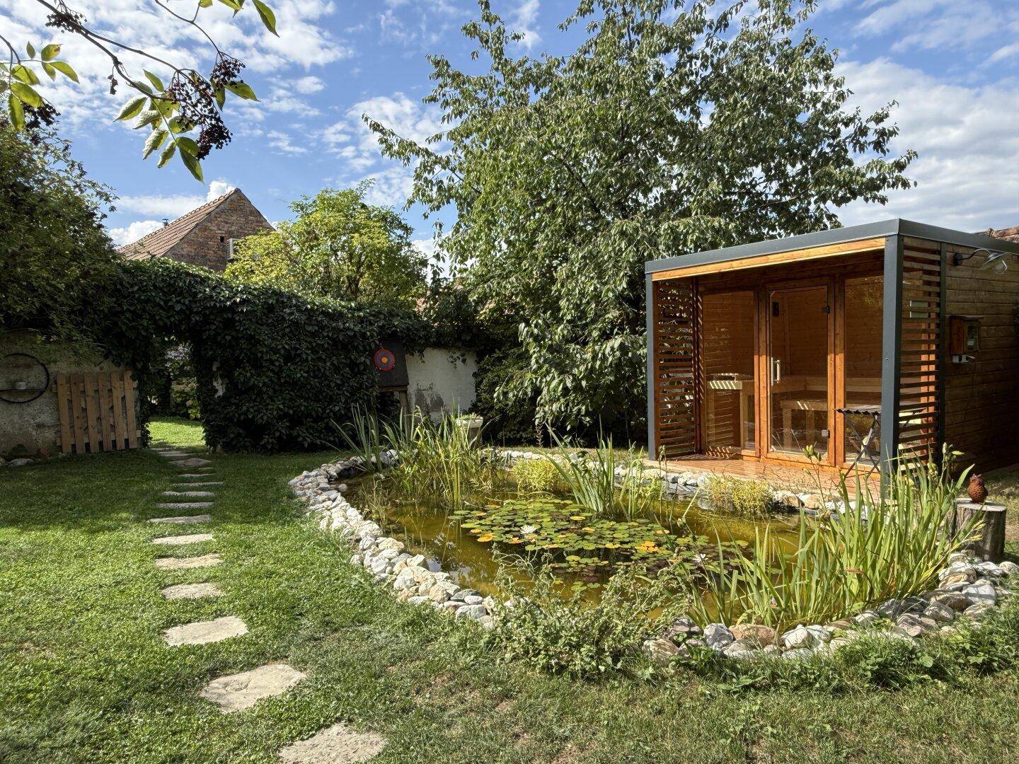 Ruhiger Gartenbereich mit moderner Sauna, Teich und Trittsteinen, umgeben von üppigem Grün.