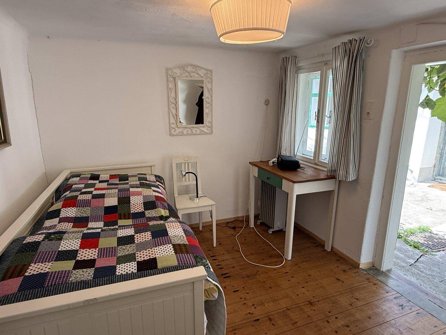 Gemütliches Schlafzimmer mit Holzboden, Einzelbett und einem kleinen Schreibtisch am Fenster.