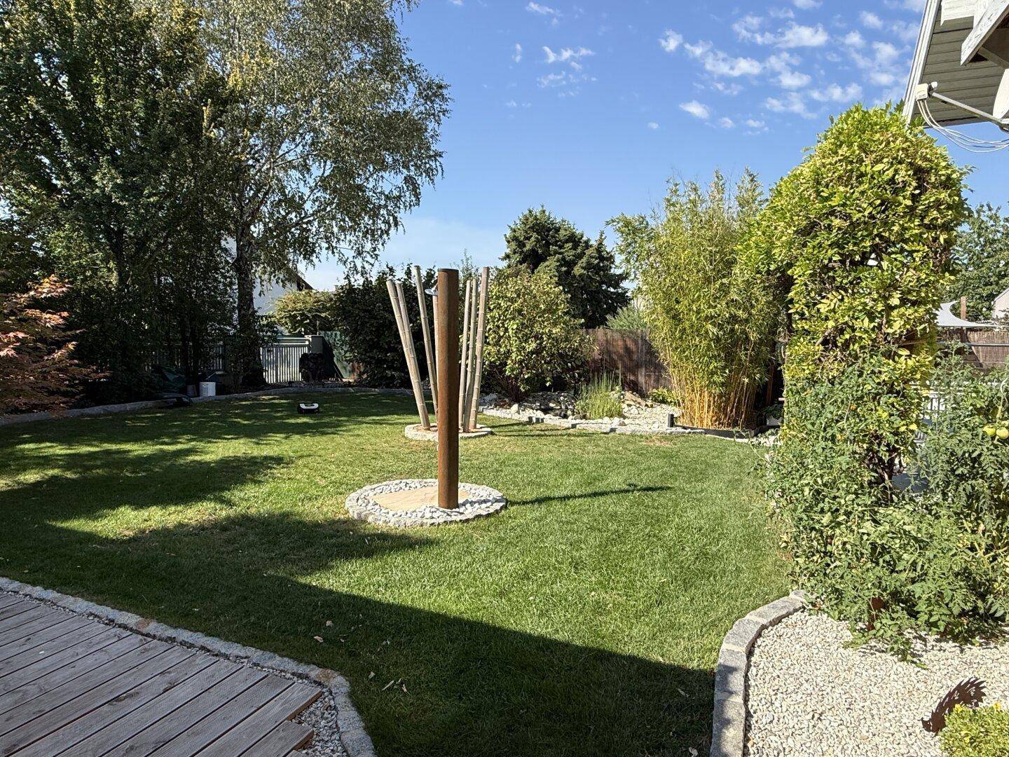 Gepflegter Garten mit Rasenfläche, Bäumen und dekorativen Elementen unter blauem Himmel.