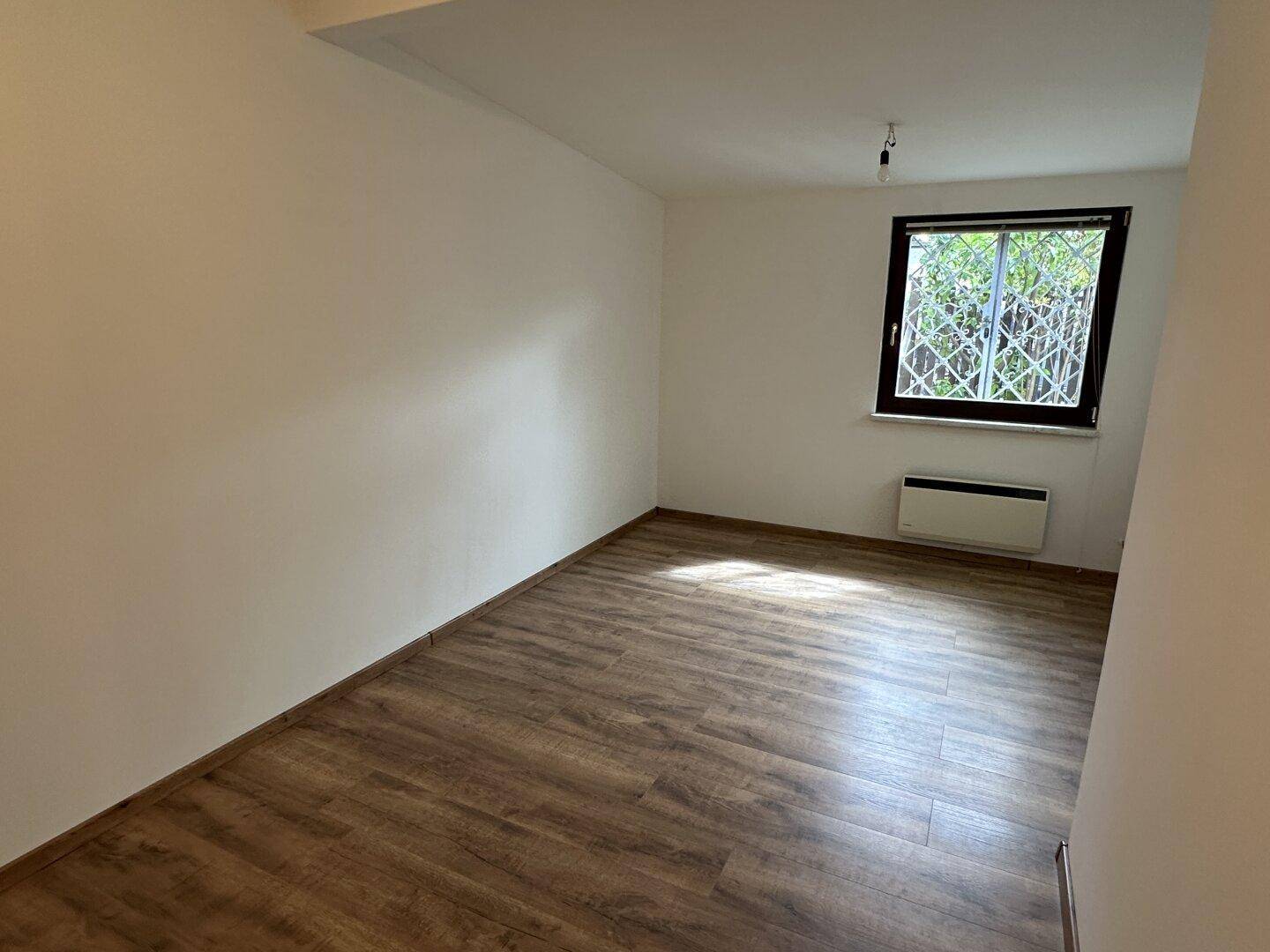Leeres Zimmer mit Laminatboden, weißen Wänden und einem Fenster mit Gitter.