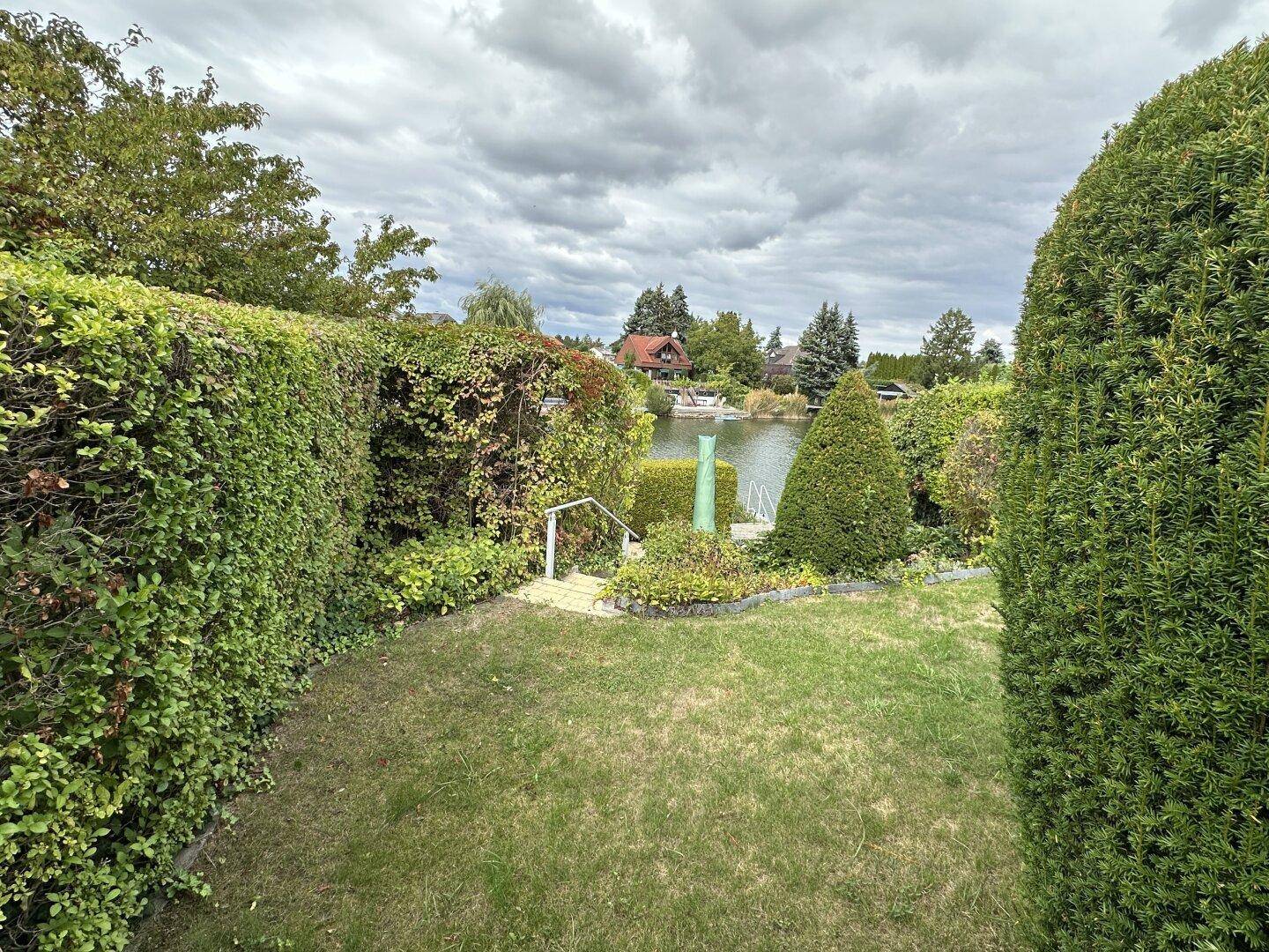 Grüner Gartenbereich mit Rasen, hohen Hecken und Zugang zum Wasser.