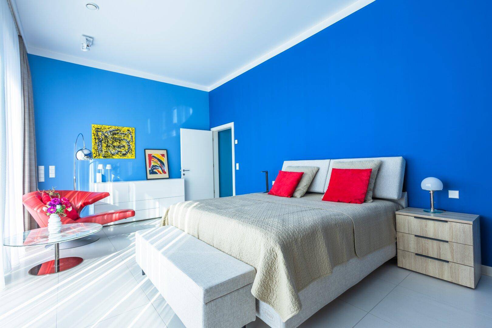 Modernes Schlafzimmer mit leuchtend blauen Wänden, großem Bett und viel Tageslicht.