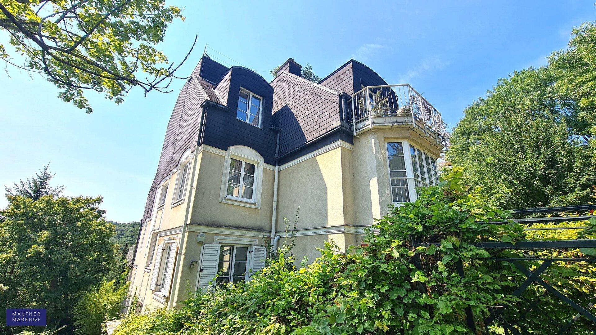 Historisches Gebäude mit Mansarddach und kleinem Balkon, eingebettet in grüne Umgebung.