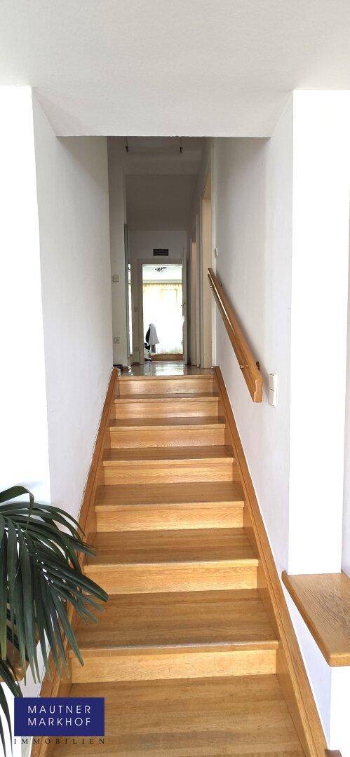 Heller Flur mit Holztreppe und schlichtem Holzgeländer, der zu weiteren Räumen führt.