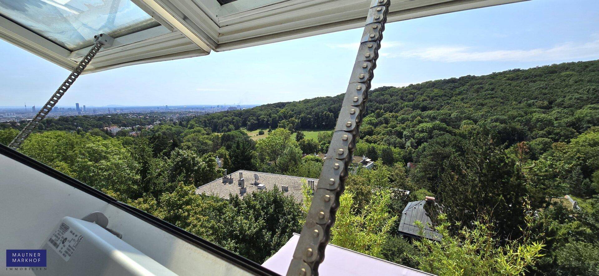Weitläufiger Blick über grüne Hügel und Wälder bis zur fernen Stadt, gesehen durch ein Dachfenster.