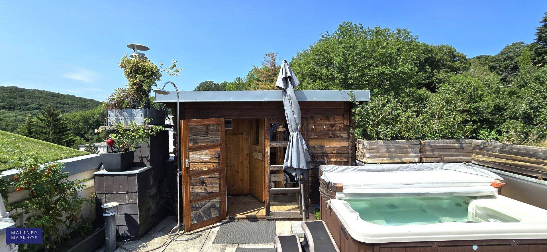 Sonniger Terrassenbereich mit Whirlpool, Saunahaus und üppiger Bepflanzung im Hintergrund.