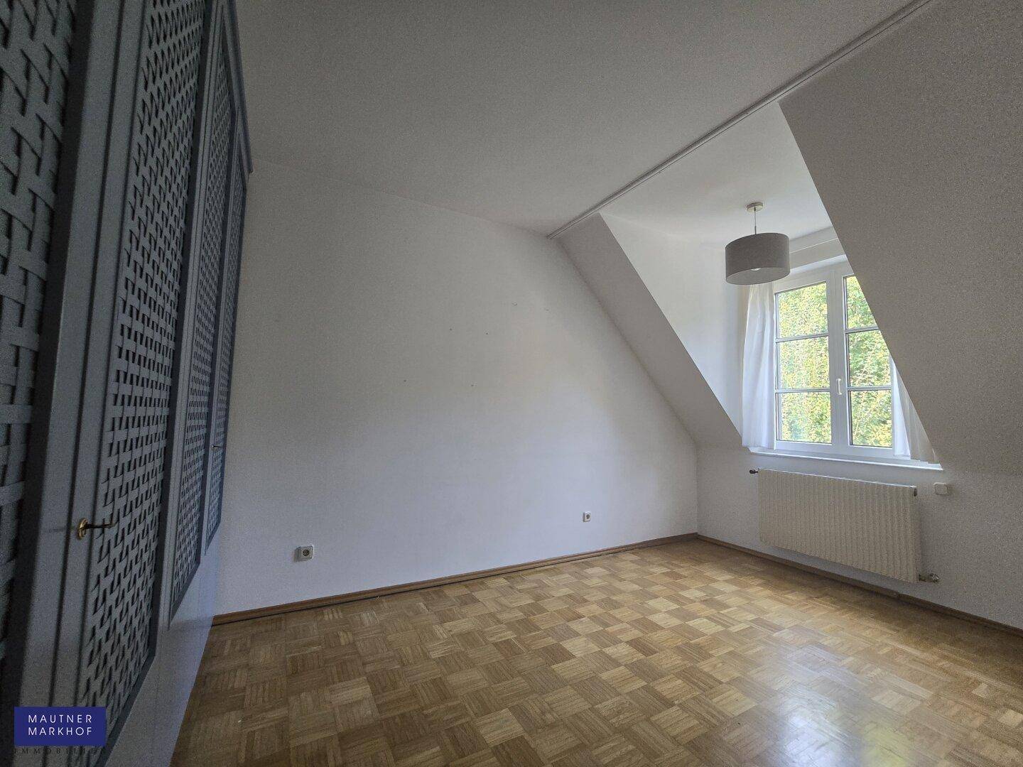 Helles Schlafzimmer mit Dachschrägen, Parkettboden und einem Fenster mit Grünblick.