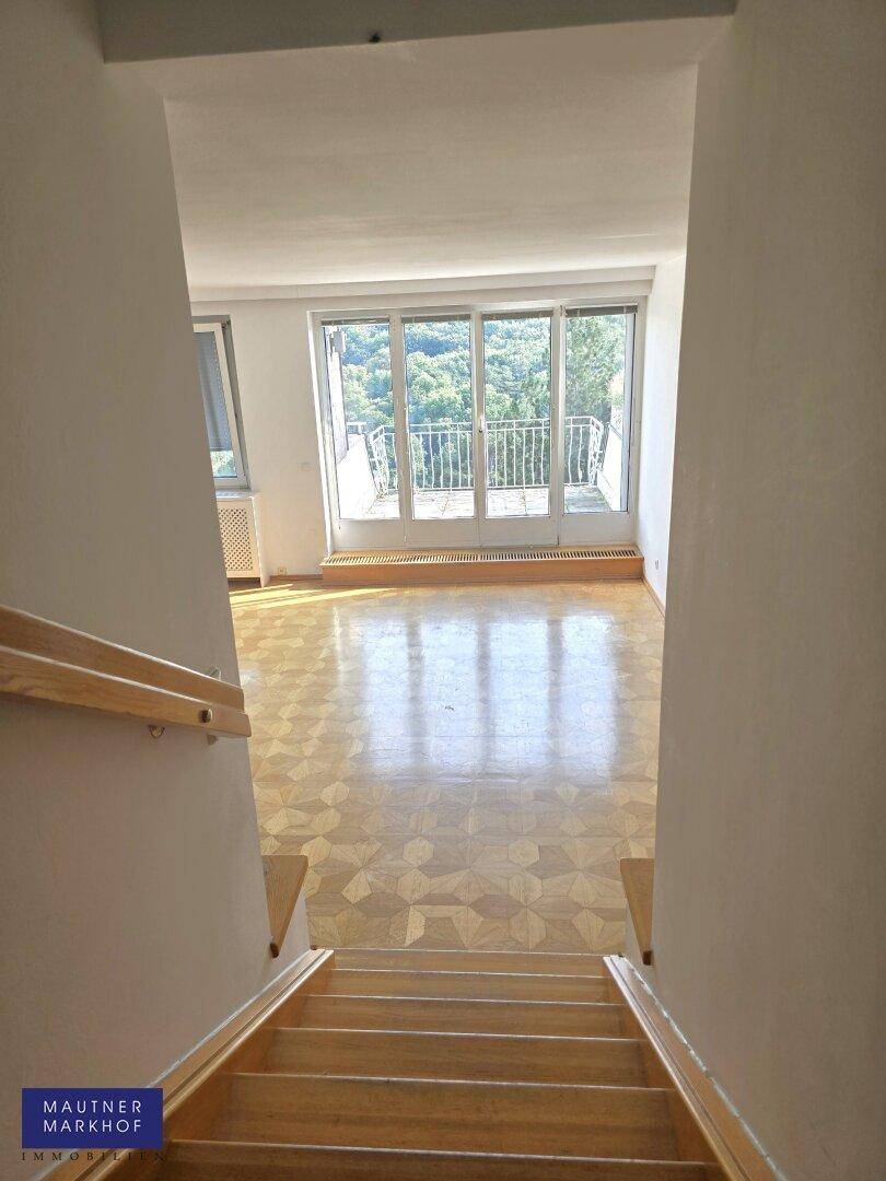 Blick von der Holztreppe in einen hellen Raum mit Parkettboden und Balkonzugang.