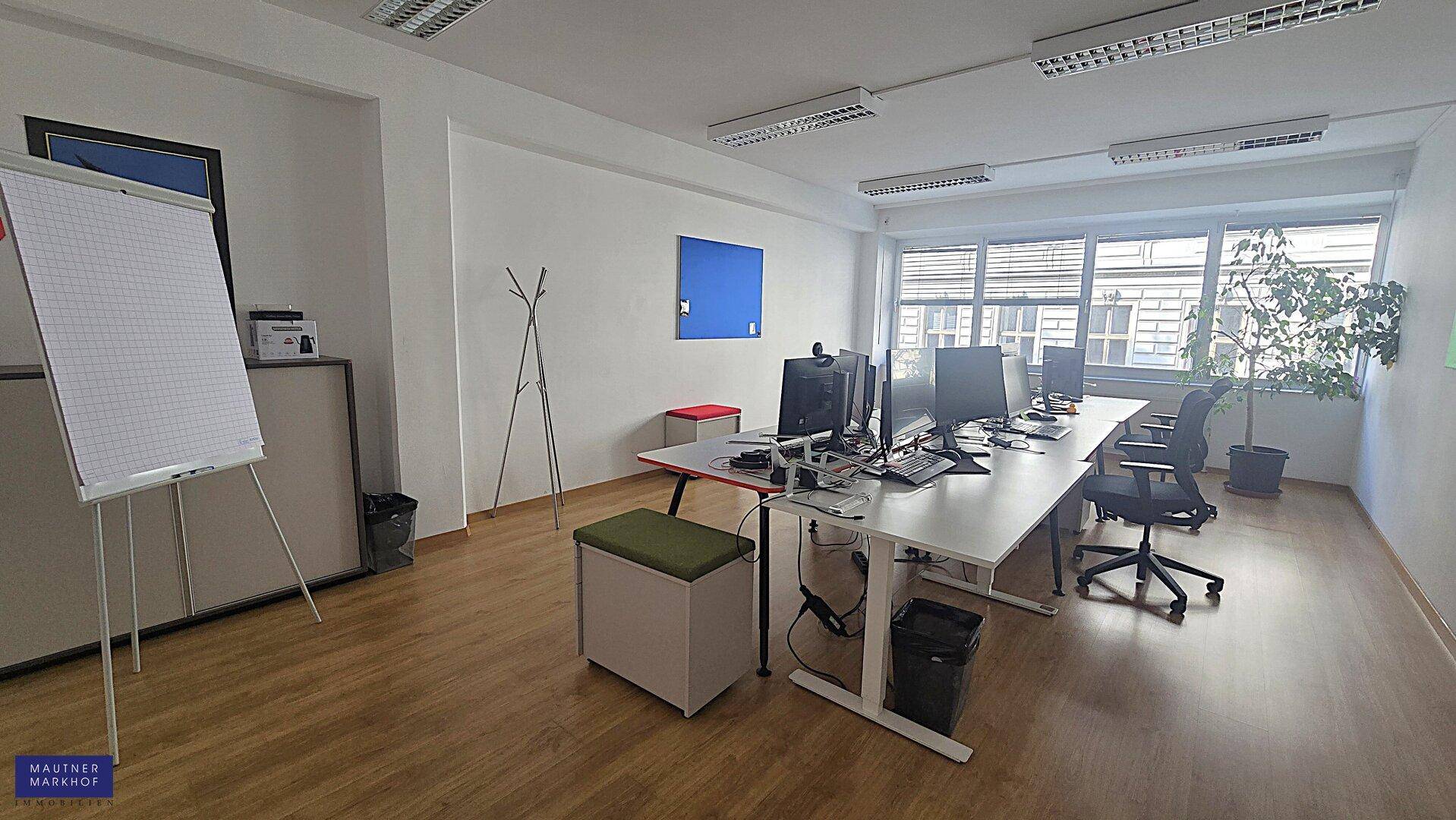 Modern eingerichtetes Büro mit mehreren Arbeitsplätzen, Computern und einem Flipchart, gut beleuchtet durch Fenster.