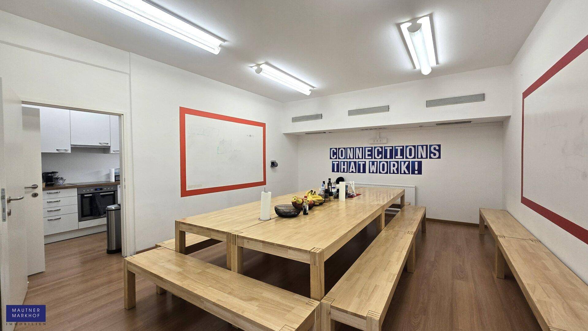 Gemeinschaftsraum mit langen Holztischen und Bänken, einer kleinen Küche im Hintergrund und inspirierenden Wanddekorationen.