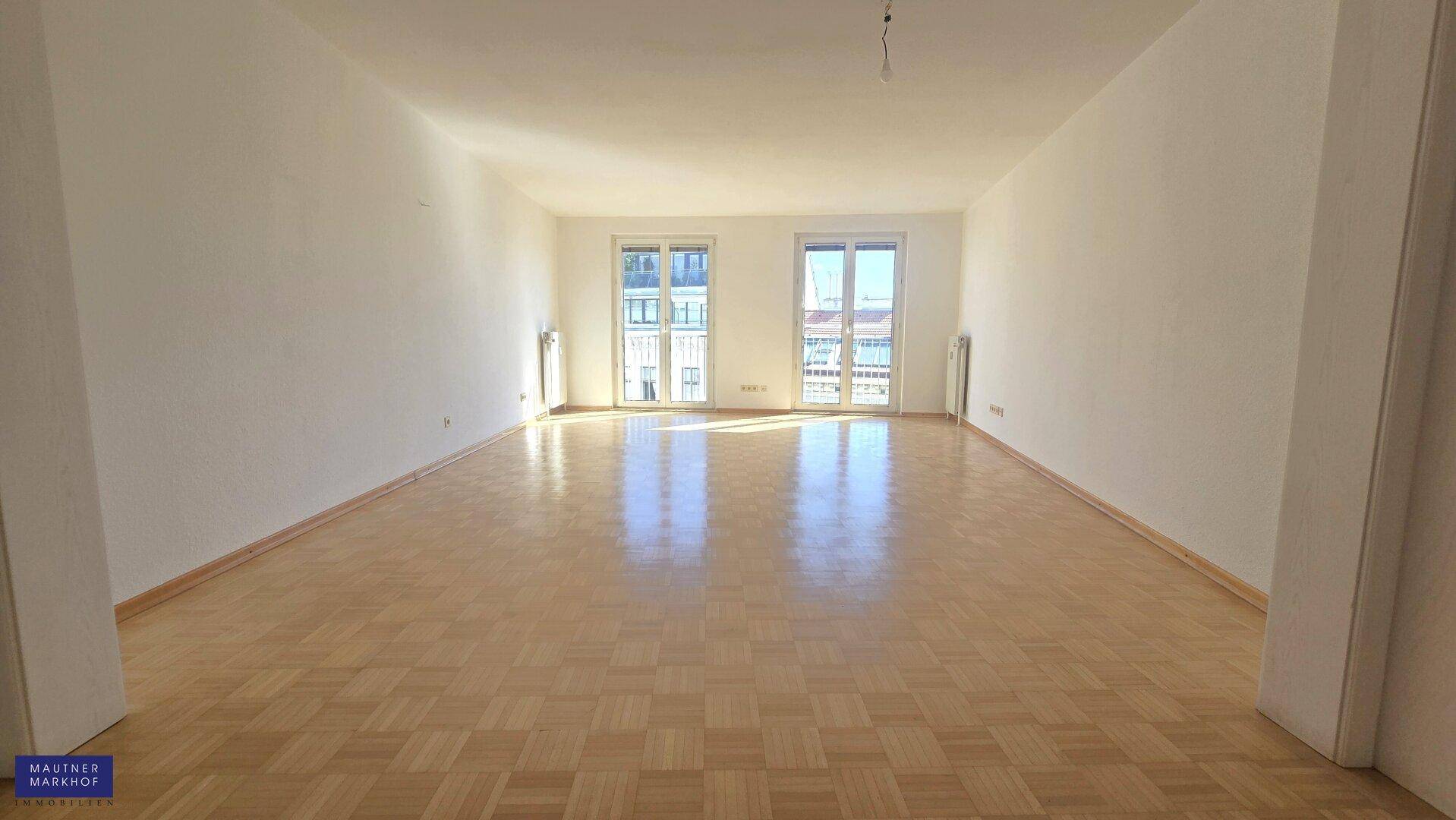 Weitläufiger Wohnraum mit hellem Parkettboden und großen Fenstern, die viel Tageslicht hereinlassen.