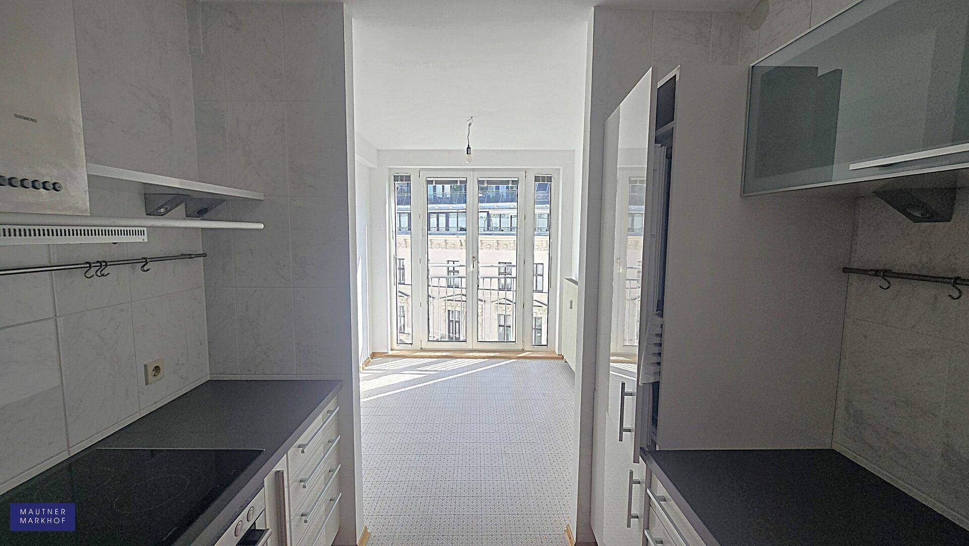 Blick aus der Küche in einen hellen Raum mit großen Fenstern und Zugang zu einem Balkon.