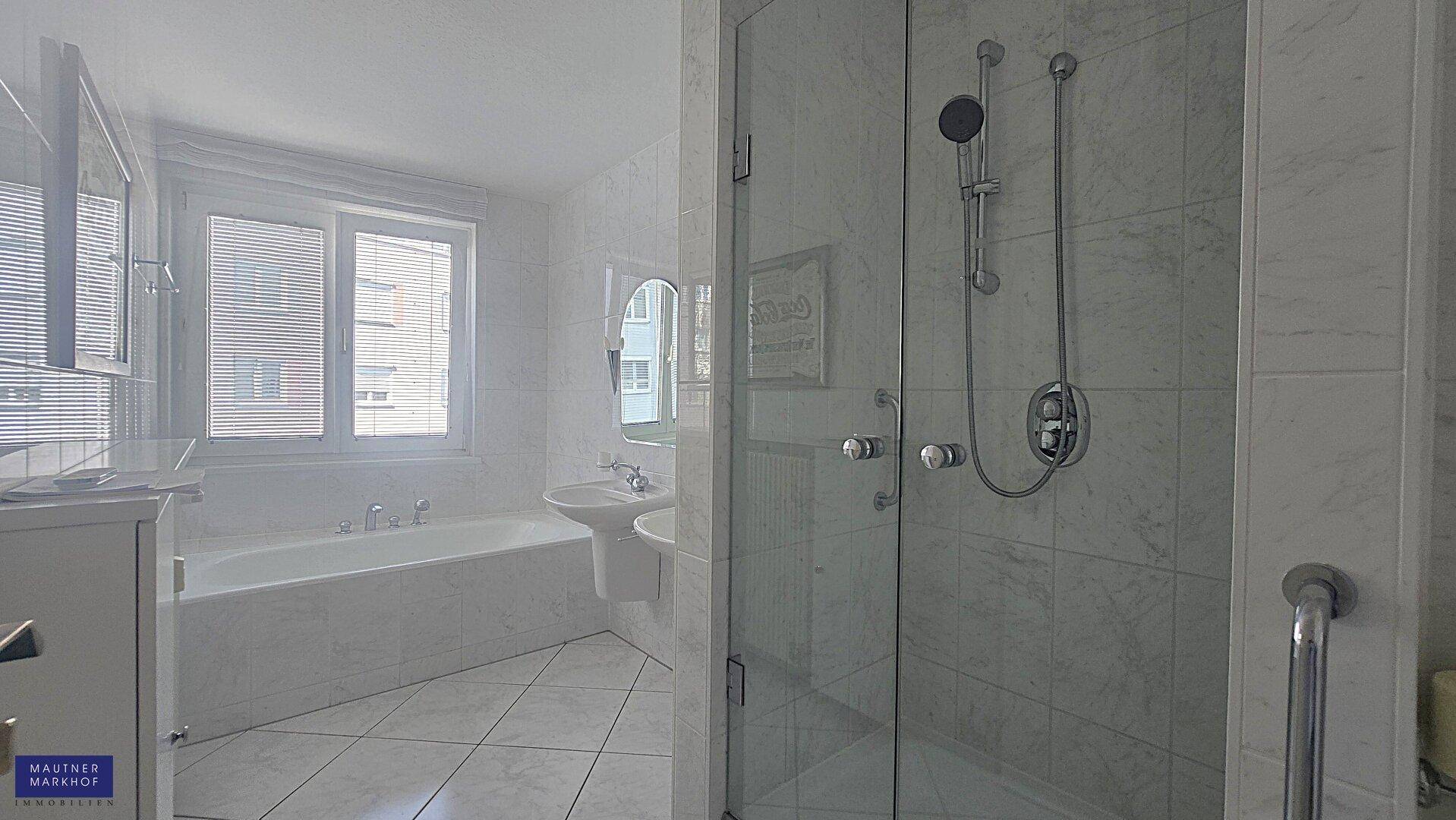 Helles Badezimmer mit weißen Fliesen, Badewanne, Duschkabine und Fenster für Tageslicht.