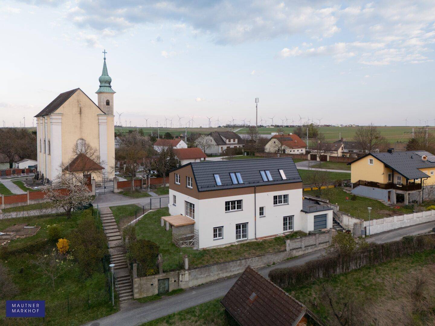Luftaufnahme eines modernen Einfamilienhauses neben einer Kirche mit Windrädern im Hintergrund.