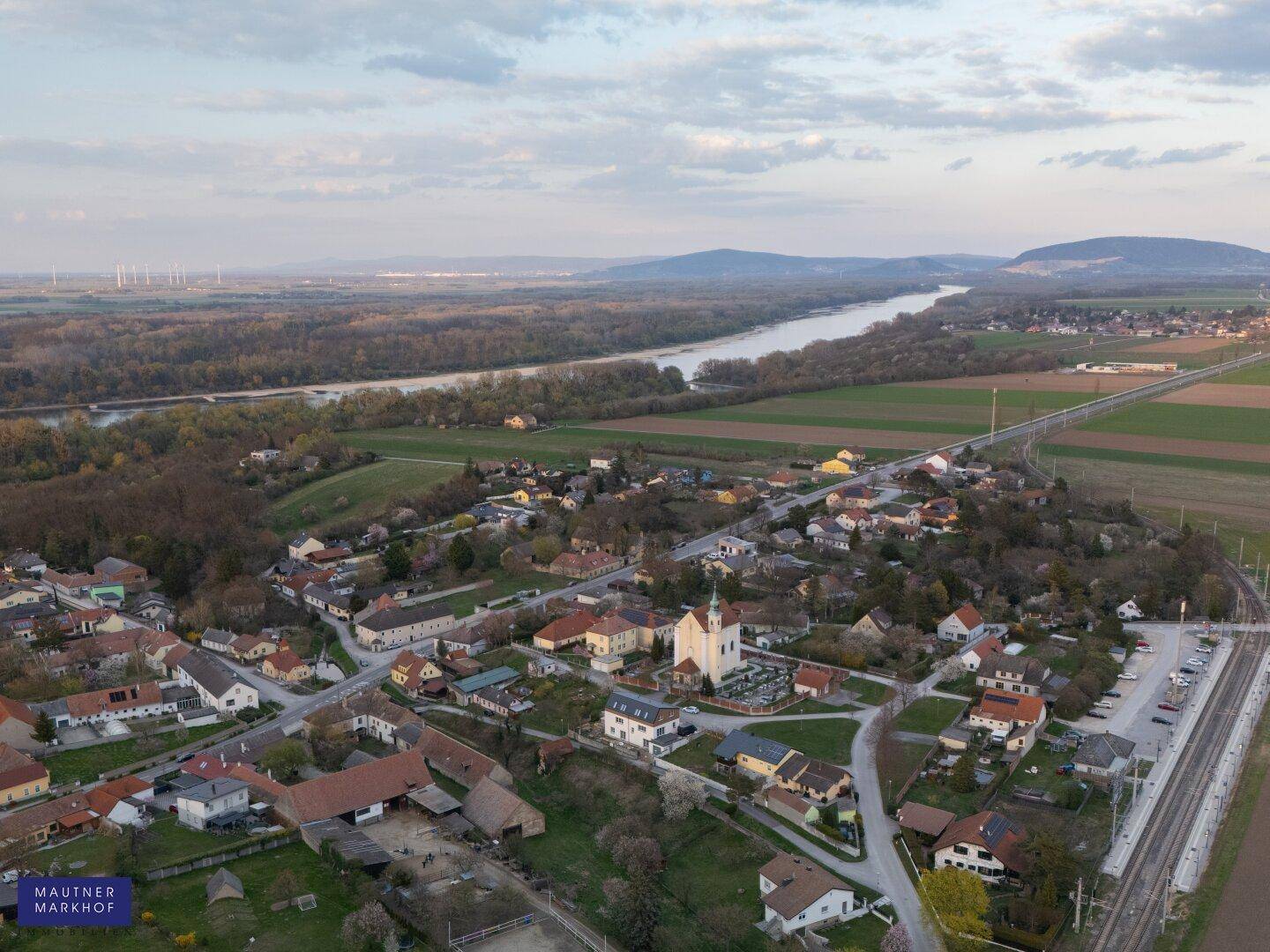 Weitläufige Luftaufnahme des Dorfes mit Fluss, Feldern und Windrädern in der Ferne.