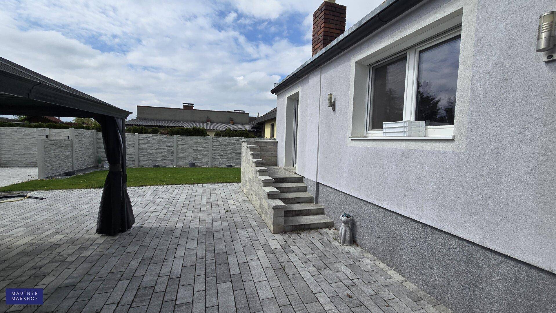 Seitenansicht des Hauses mit gepflastertem Bereich, Außentreppe und Garten.