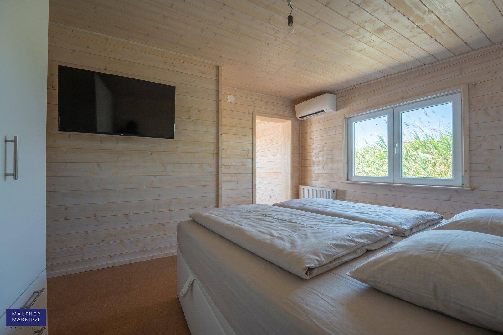 Schlafzimmer mit Holzwänden, Flachbildfernseher und Fenster mit Blick ins Grüne.