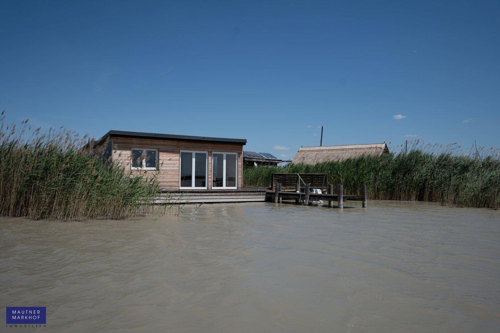 Holzhaus am Seeufer, umgeben von Schilf, mit einem weiteren Gebäude im Hintergrund.