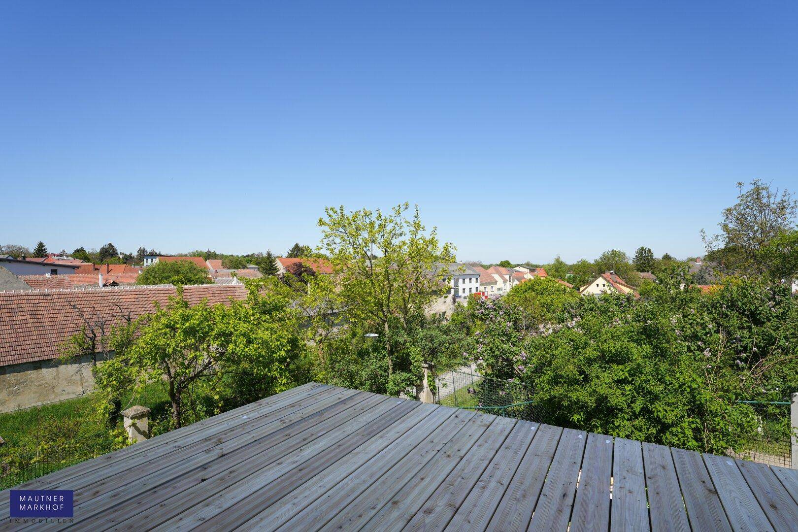 Weitläufiger Blick von einer Holzterrasse über die grüne Wohngegend unter blauem Himmel.