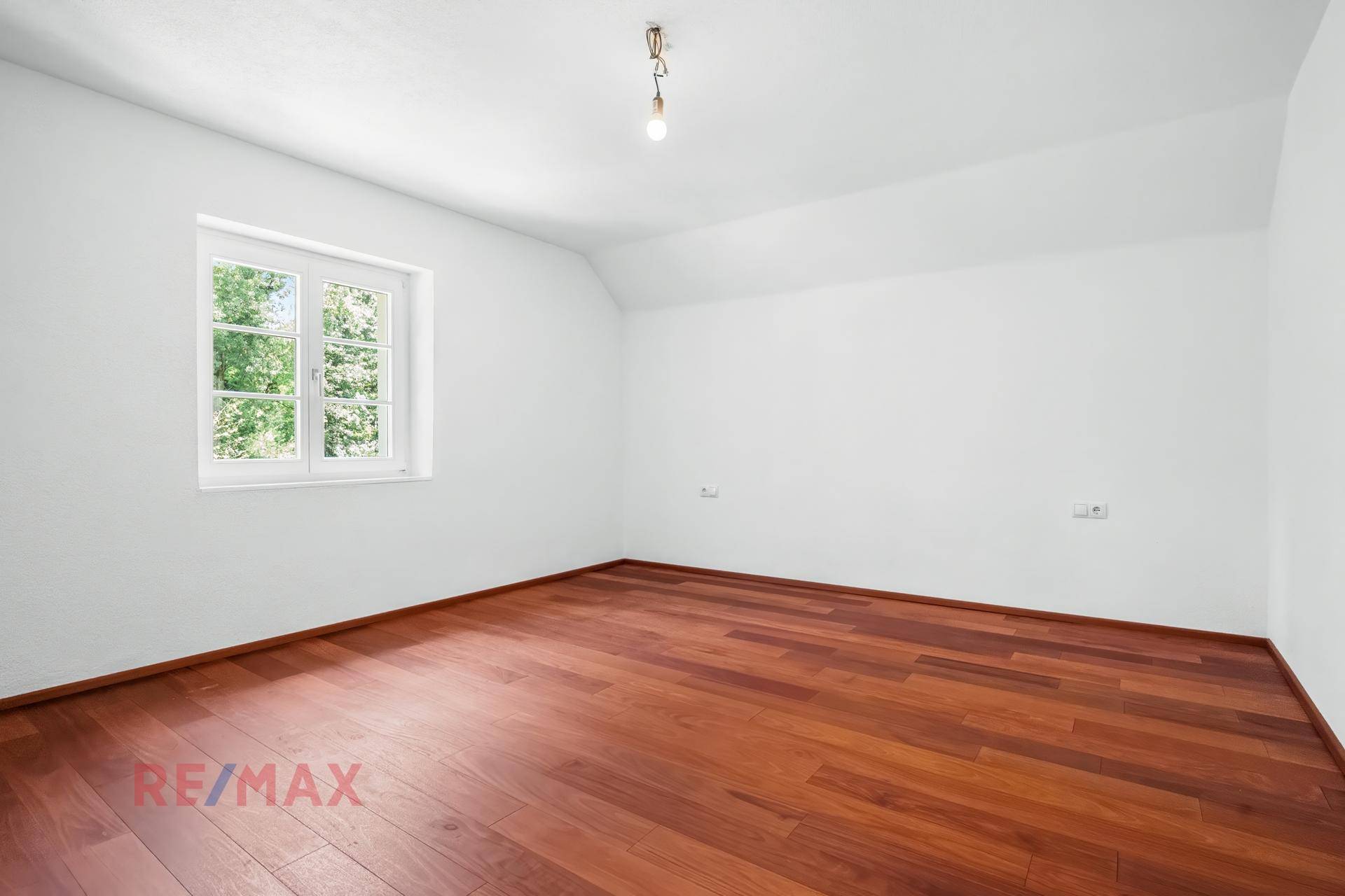 Leeres Zimmer mit Holzboden und einem Fenster, das viel Tageslicht hereinlässt und einen grünen Ausblick bietet.