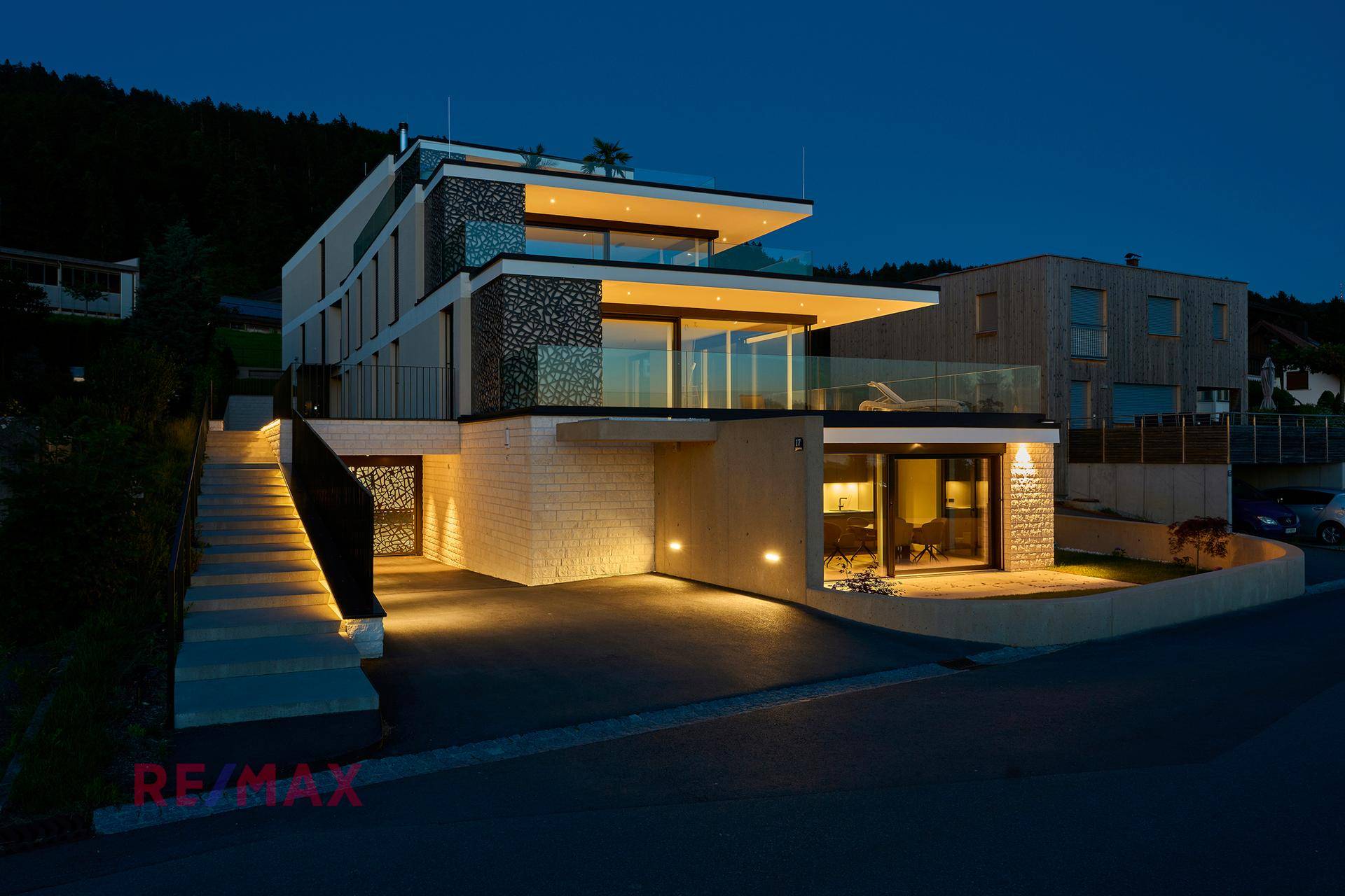 Beleuchtetes modernes Wohngebäude bei Nacht mit Terrassen und Tiefgaragenzufahrt.