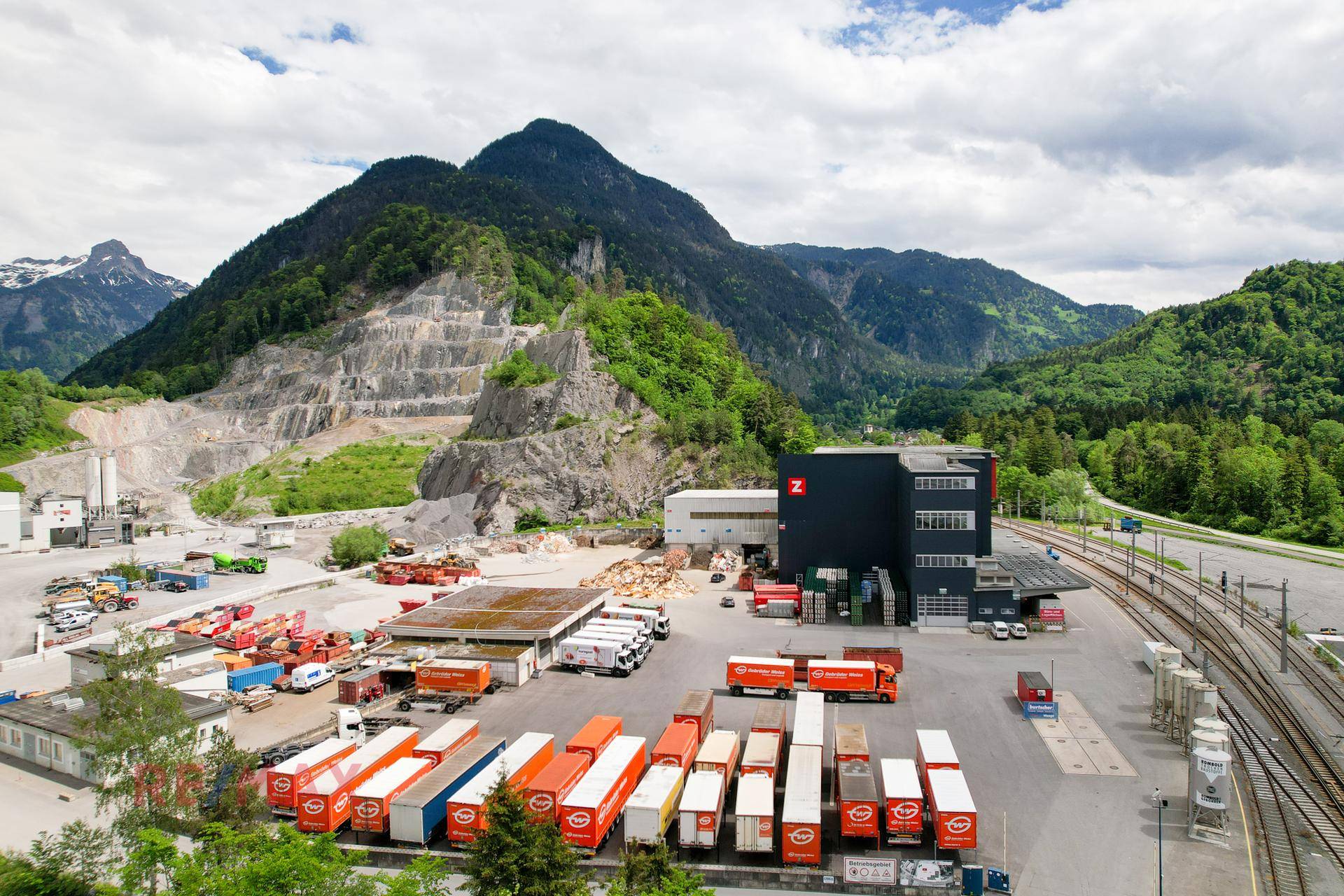 Weitläufiges Industrieareal mit Lagerflächen, Güterzügen und beeindruckender Bergkulisse unter bewölktem Himmel.