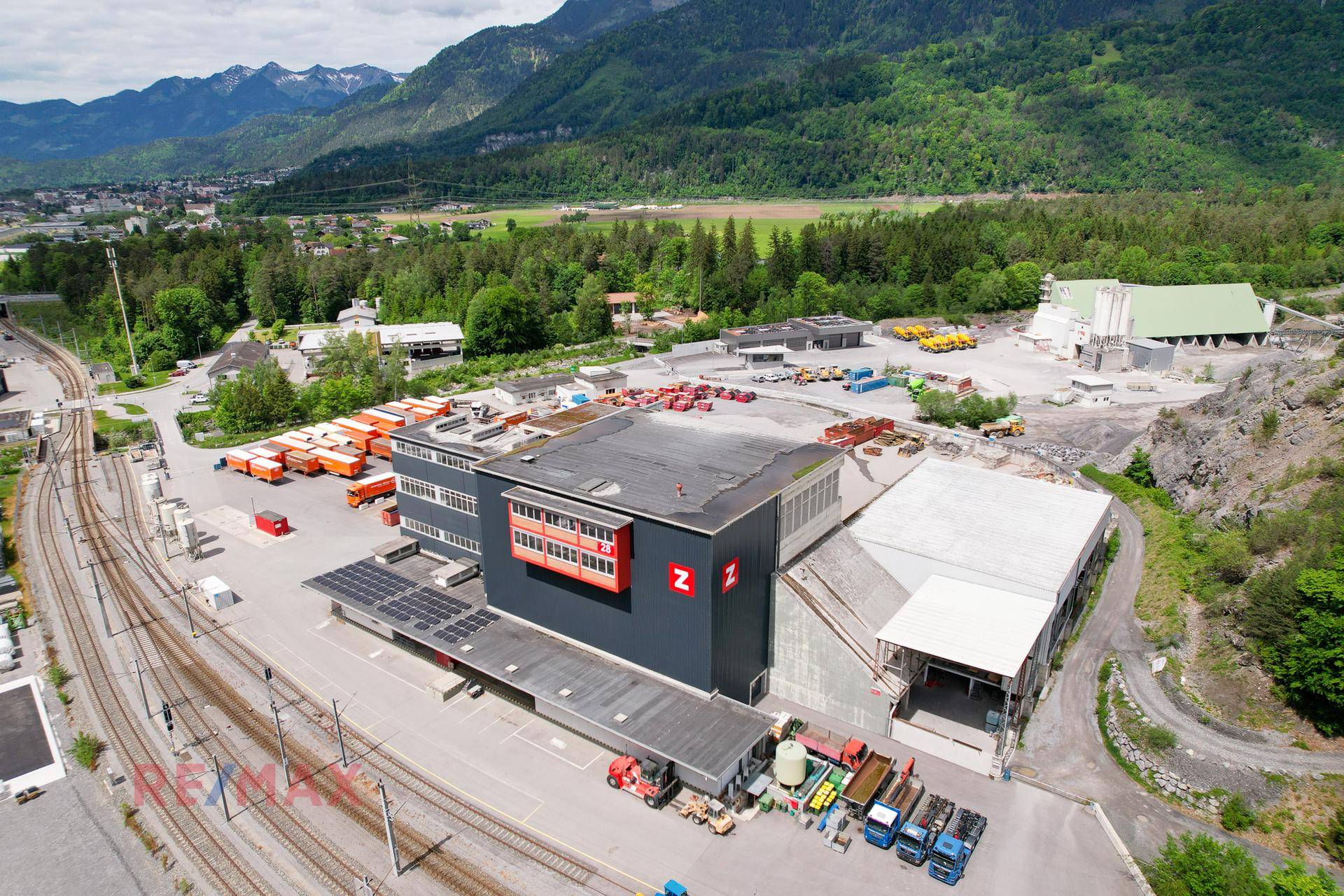 Überblick über das Industriegebäude mit angrenzenden Lagerbereichen und Bahngleisen vor einer grünen Berglandschaft.