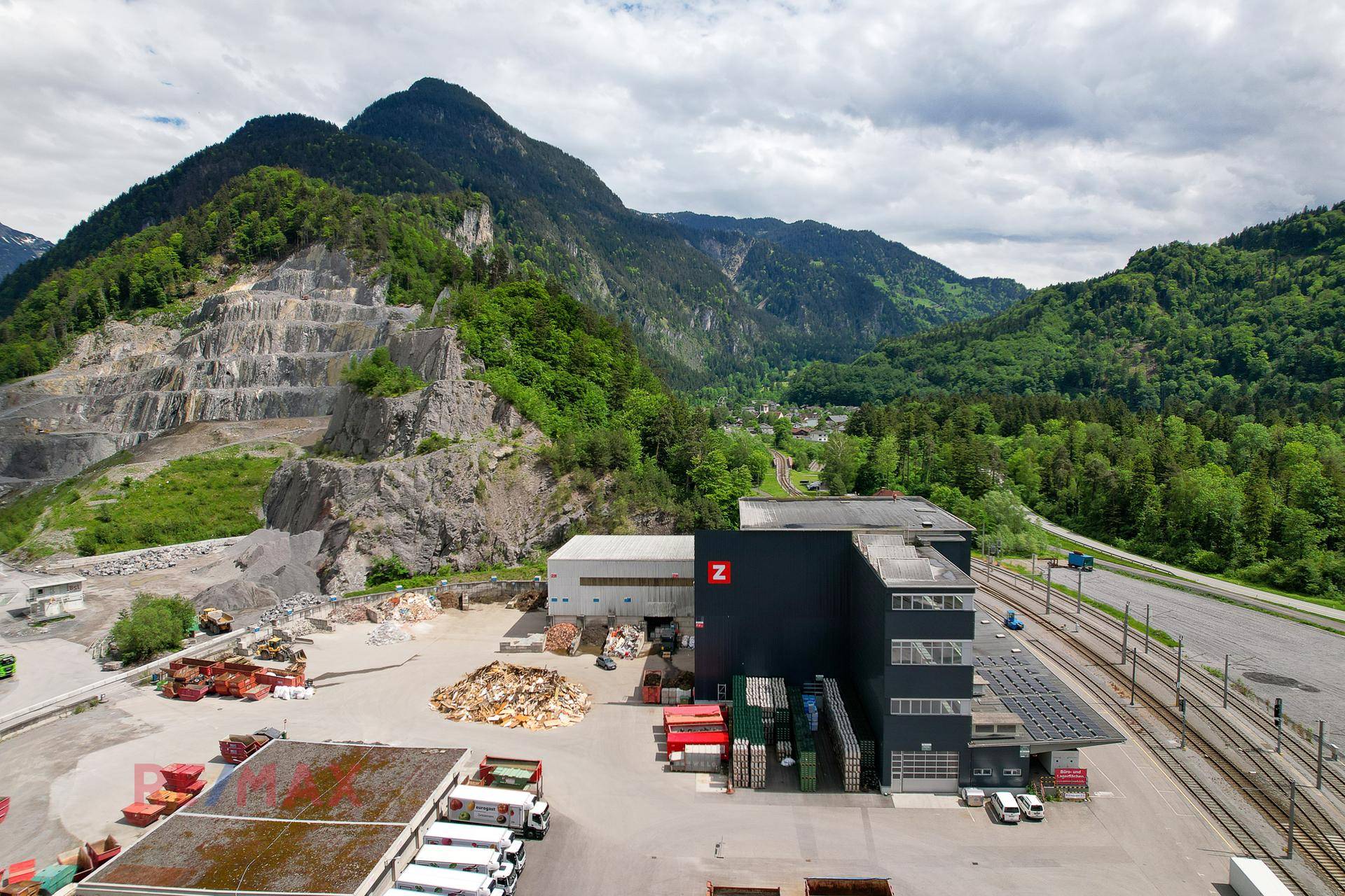 Industrielle Anlage mit Lagerhallen und Bahngleisen, eingebettet in eine grüne Berglandschaft mit Steinbruch.