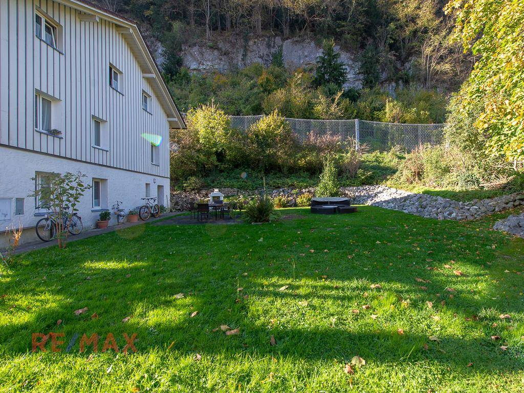 Sonniger Gartenbereich mit Rasen und Blick auf die bewaldete Hanglage hinter dem Haus.