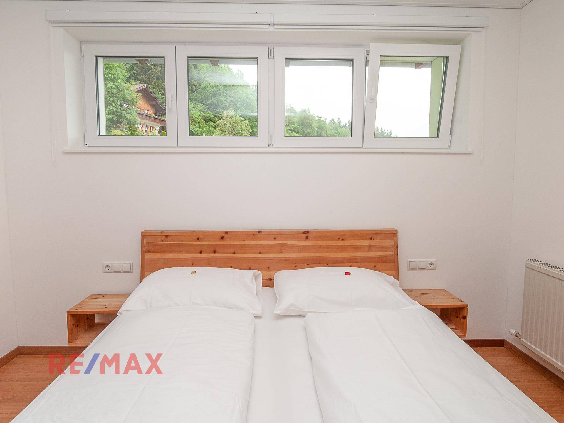 Schlafzimmer mit Doppelbett und Holzrahmen, unter einem breiten Fenster, das Tageslicht hereinlässt.