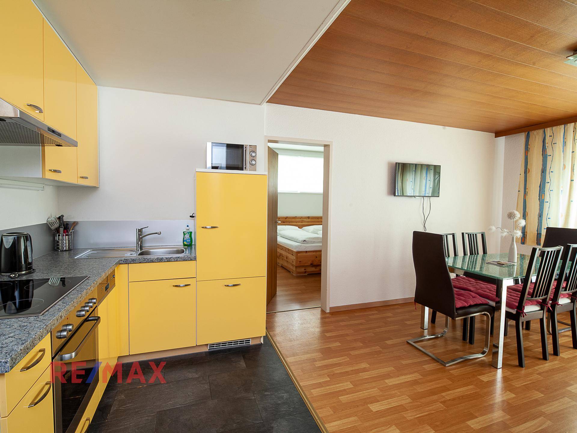 Offener Wohnbereich mit gelber Küchenzeile, Esstisch und Zugang zu einem Schlafzimmer.