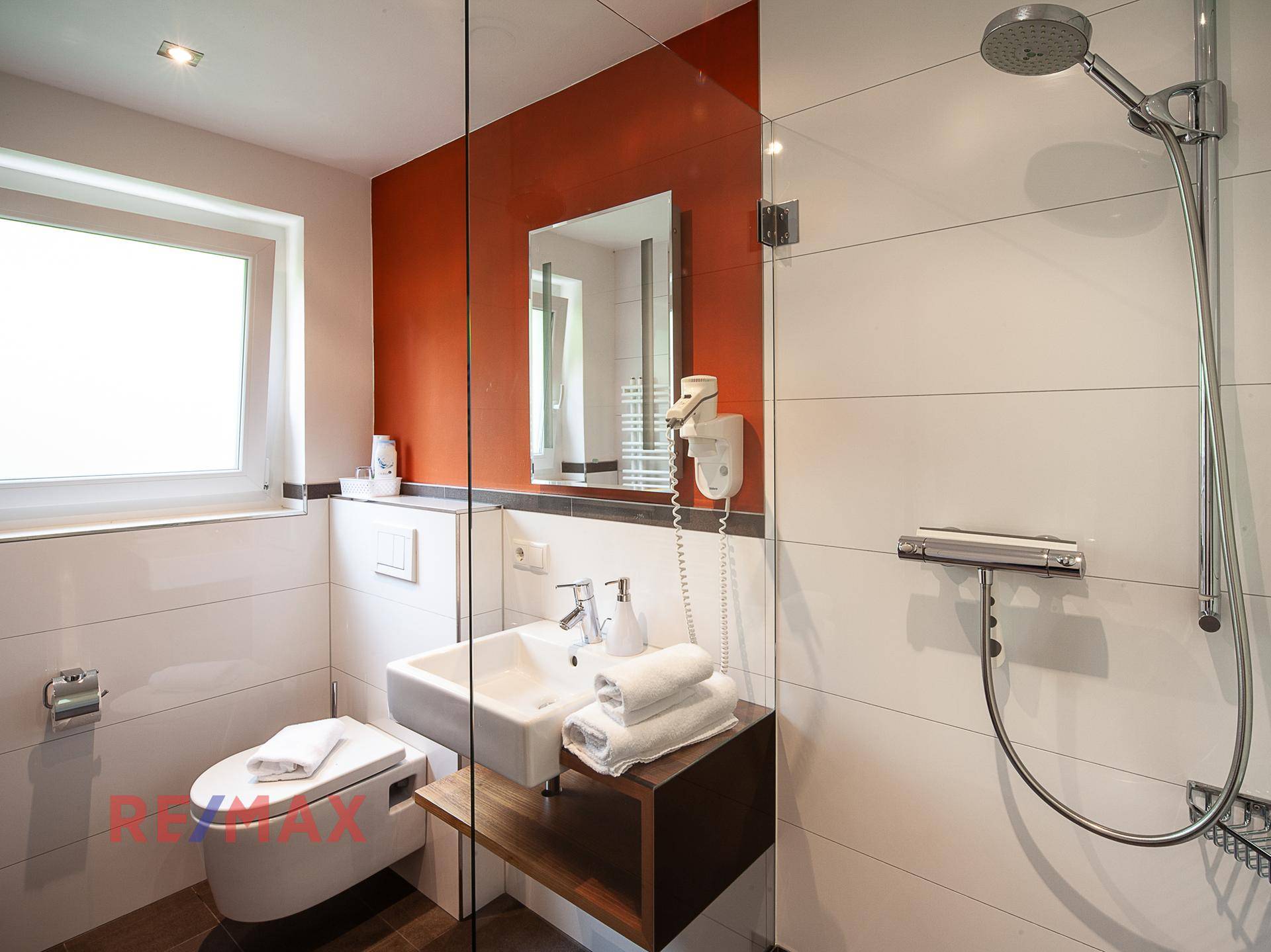 Modernes Badezimmer mit Duschkabine, Waschbecken und Fenster, in Weiß und Orange gehalten.