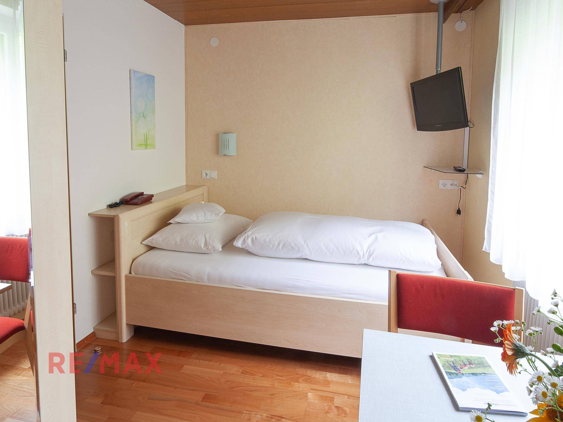 Einzelzimmer mit Holzbett, Schreibtisch und Wandfernseher, gut beleuchtet durch ein Fenster.