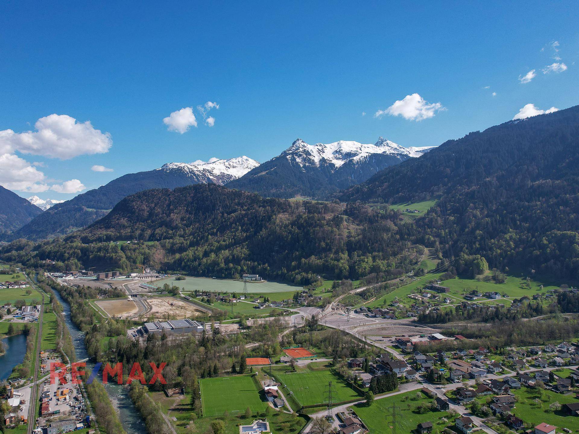 Weitwinkel-Luftaufnahme der alpinen Landschaft mit See, schneebedeckten Bergen und grünen Feldern.