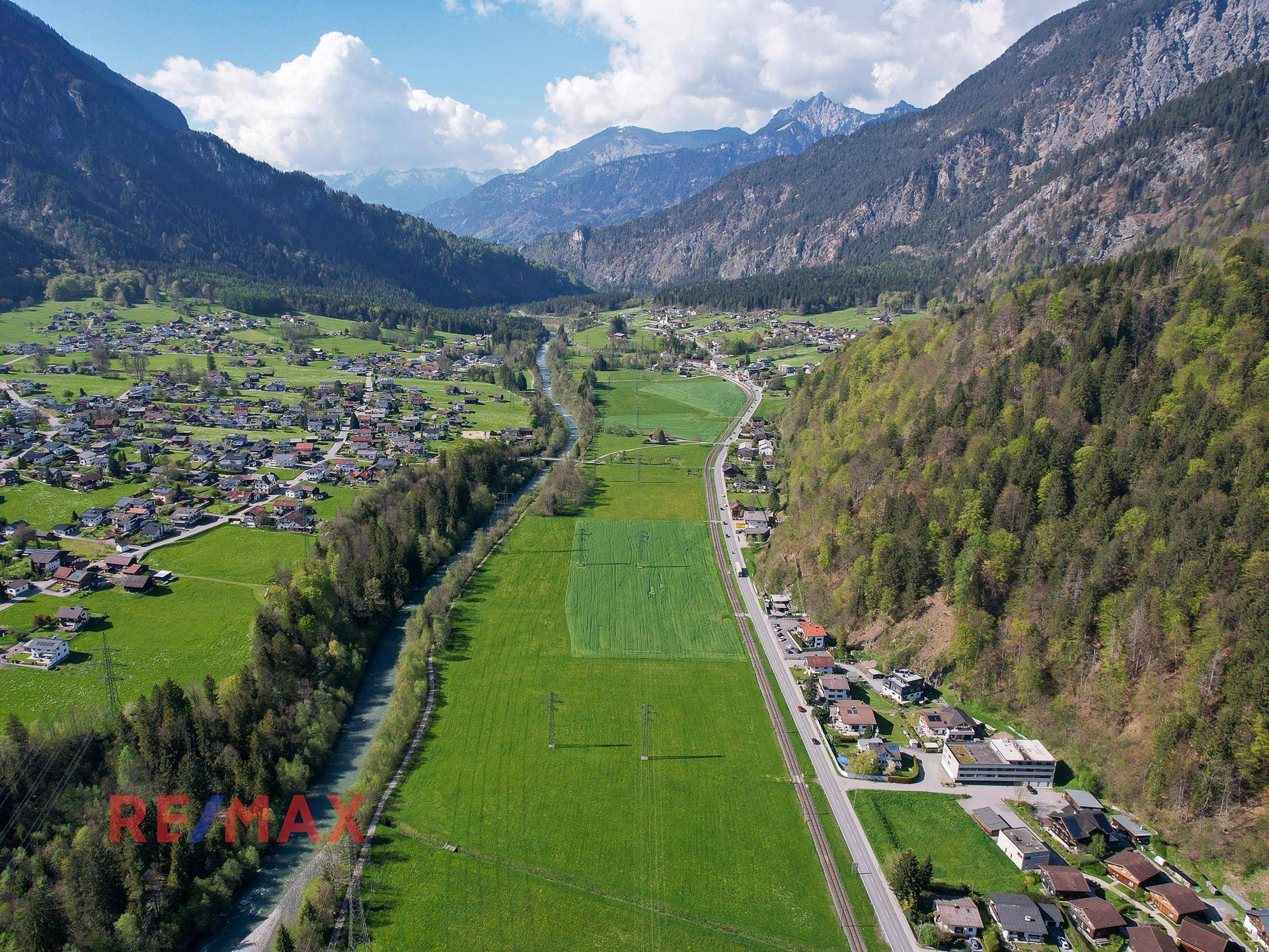 Panoramablick auf ein grünes Tal mit Fluss, Feldern, Straße und Siedlungen, umgeben von Bergen.