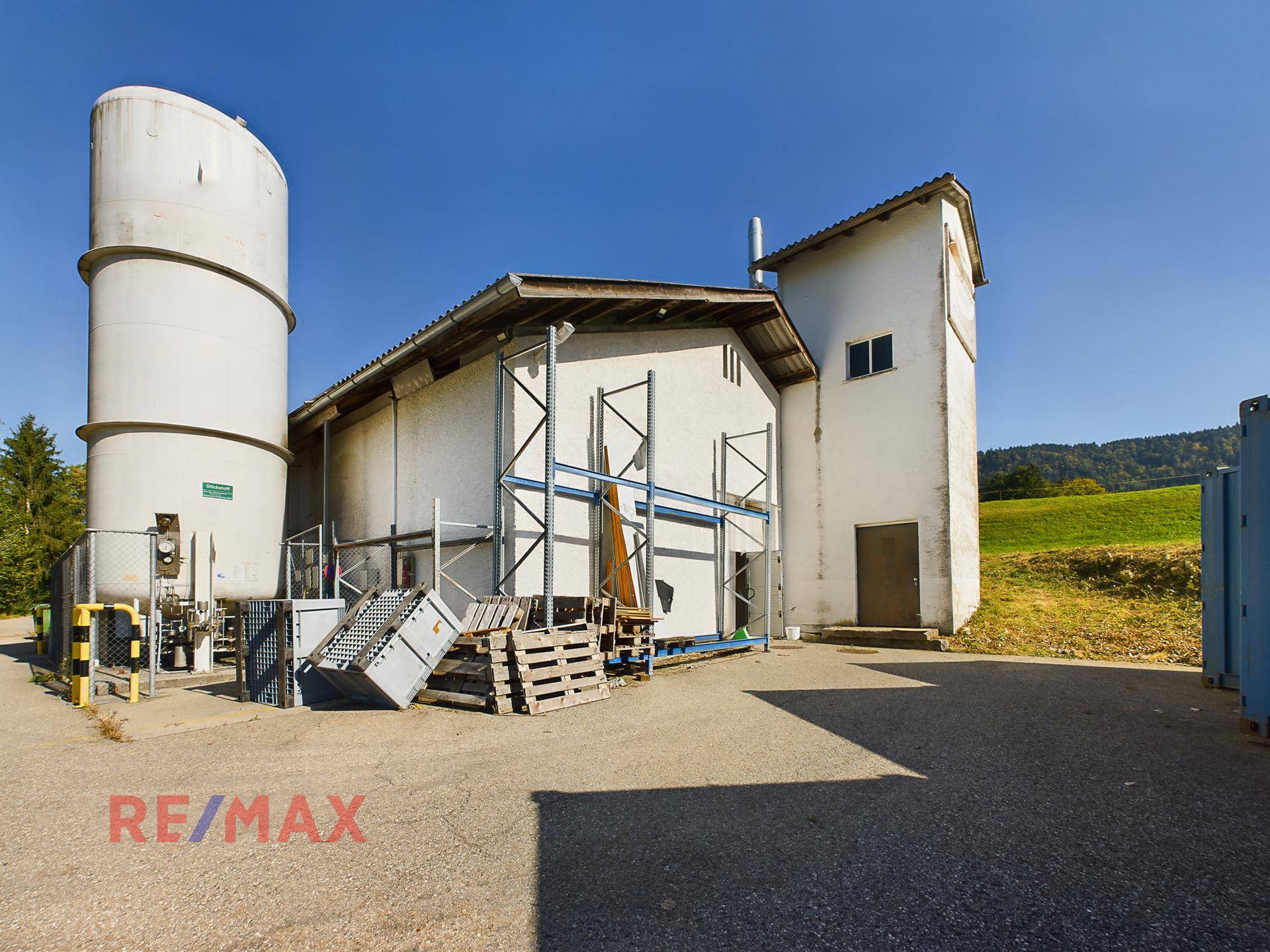 Außenansicht eines Gewerbegebäudes mit großem Silo und umgebender Grünfläche unter blauem Himmel.
