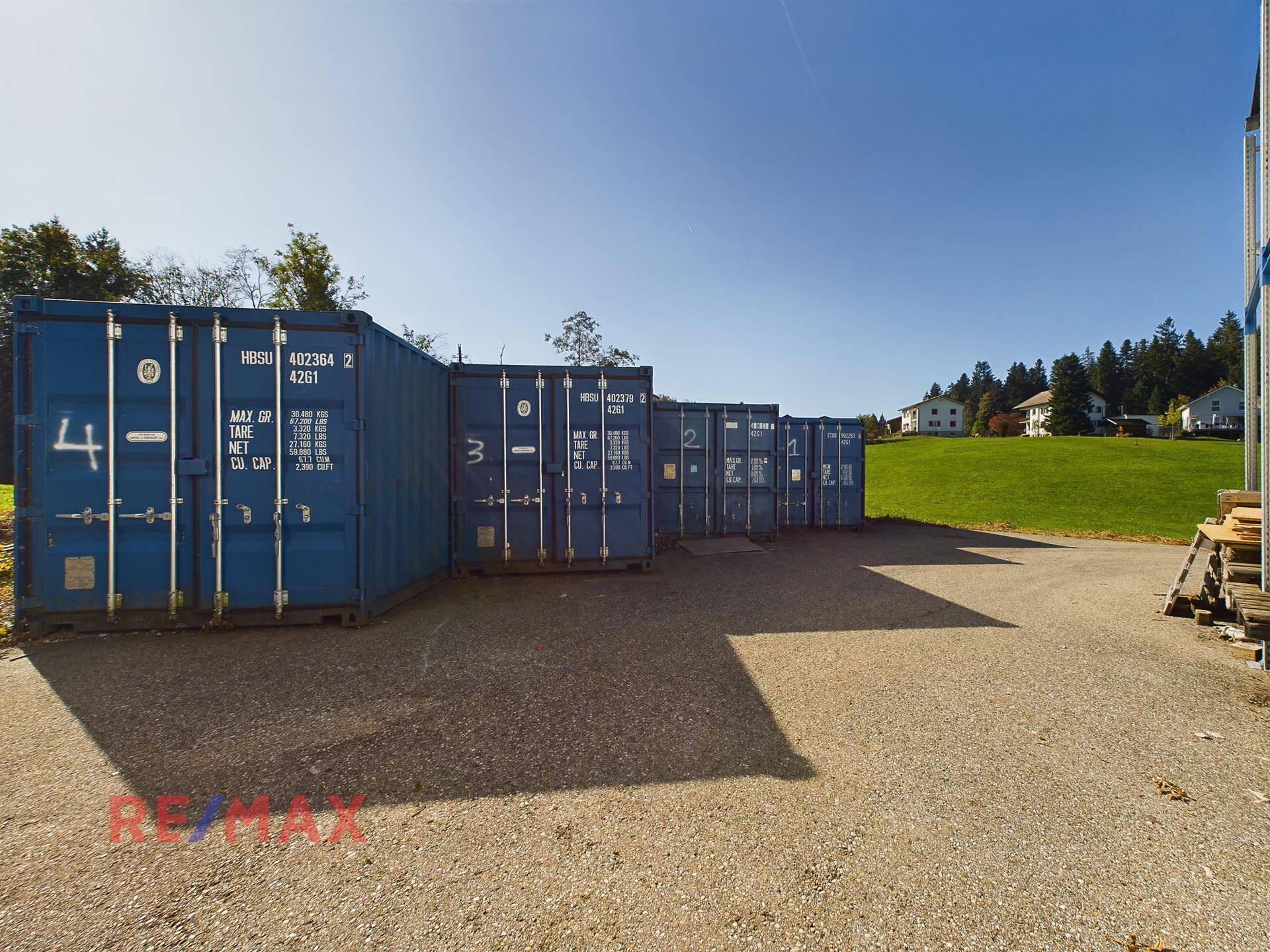 Reihe blauer Lagercontainer auf einem Schotterplatz neben einer grünen Wiese unter blauem Himmel.