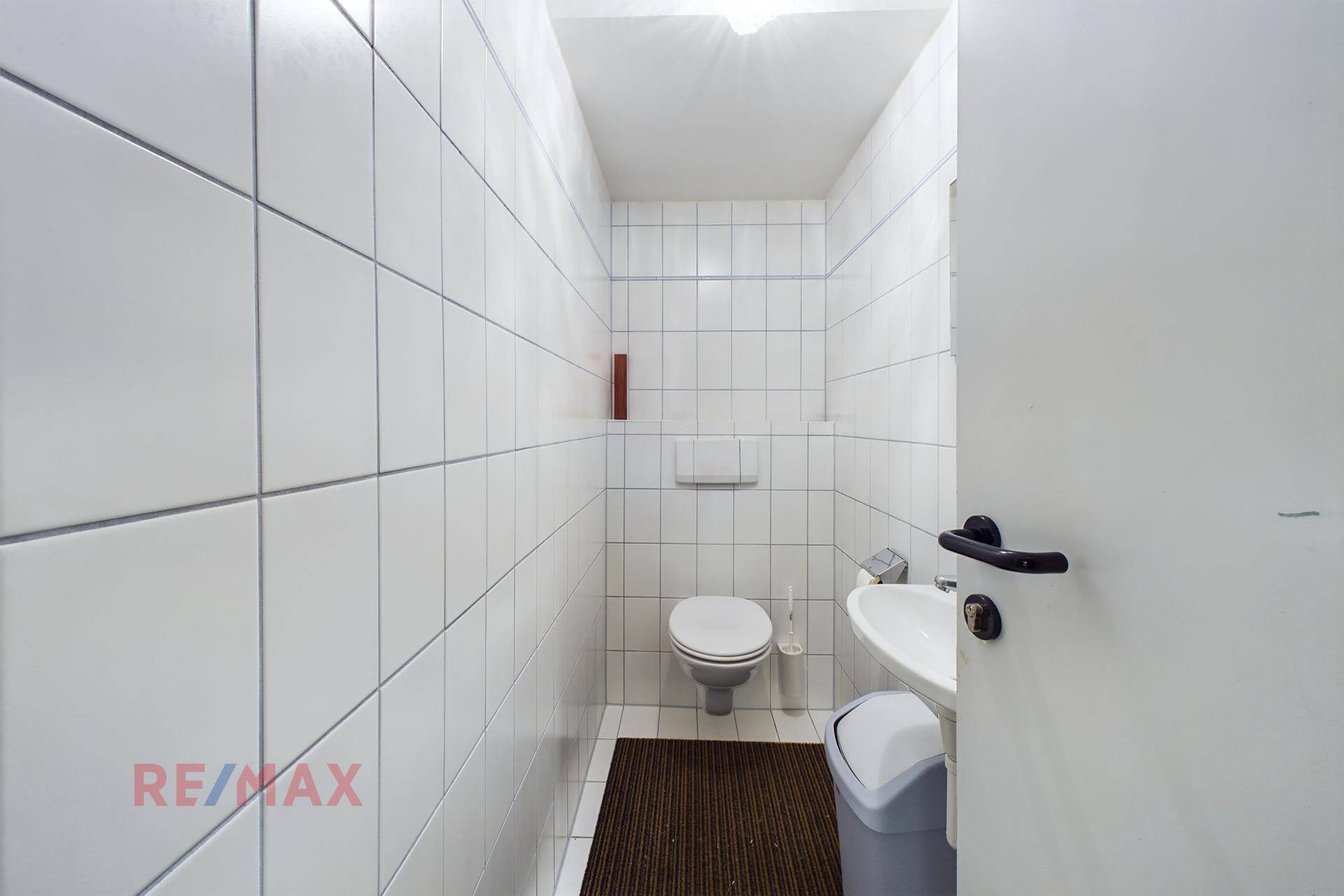 Einfache Toilette mit weißen Fliesen, WC, Waschbecken und einem kleinen Mülleimer.