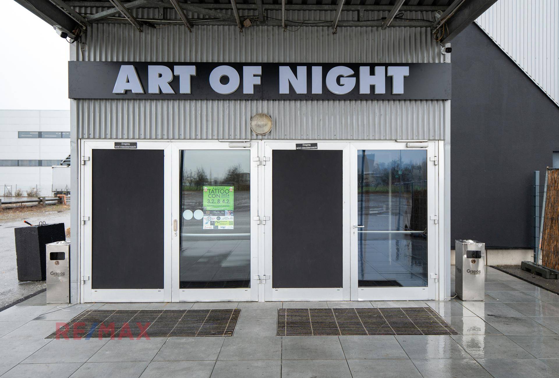 Eingangsbereich des Gebäudes 'ART OF NIGHT' mit Glastüren und Vordach an einem bewölkten Tag.