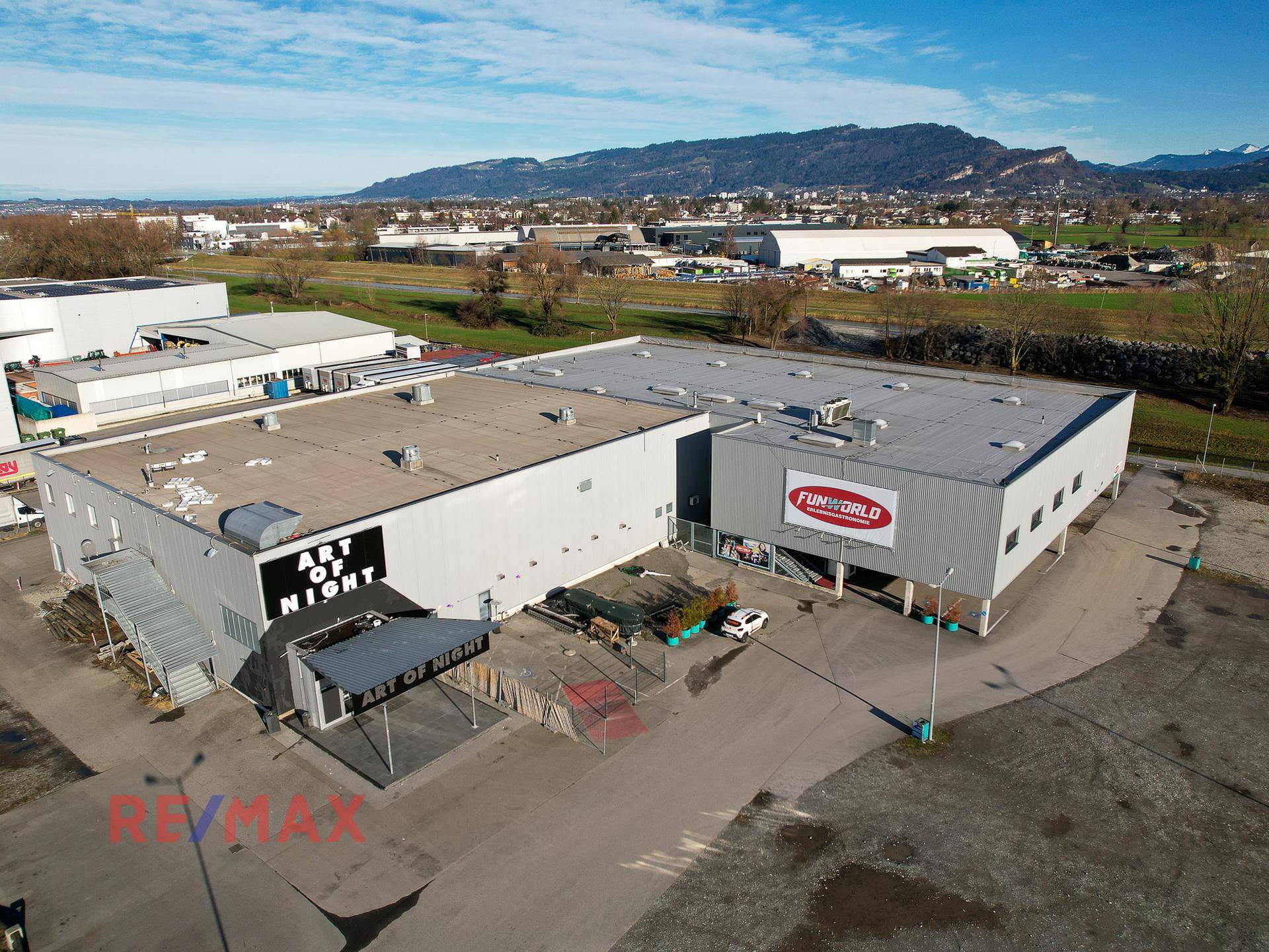 Luftaufnahme des Gewerbegebiets mit Gebäuden, Parkplätzen und Bergen im Hintergrund unter blauem Himmel.